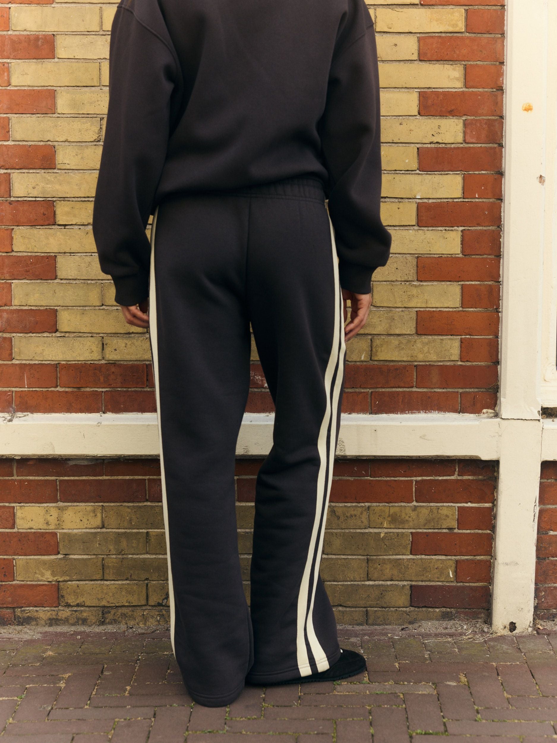 Rear view of a woman wearing espresso straight-leg sweatpants with side piping against a brick wall.