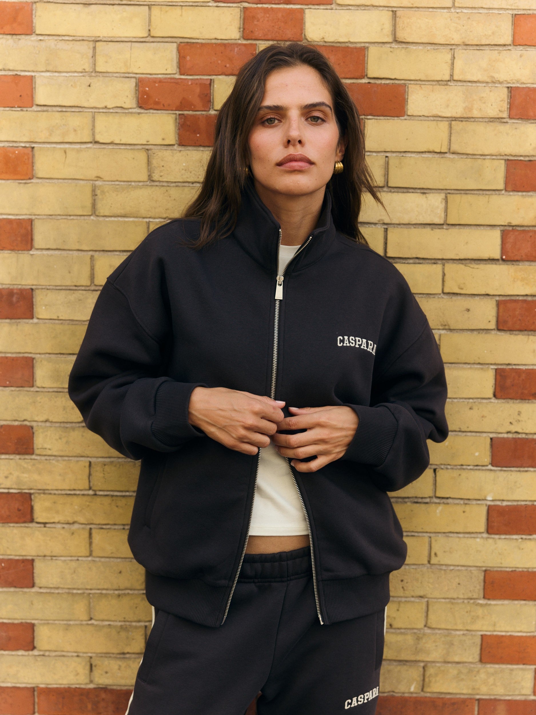 Woman wearing an espresso brown funnel zip jacket against a brick wall.