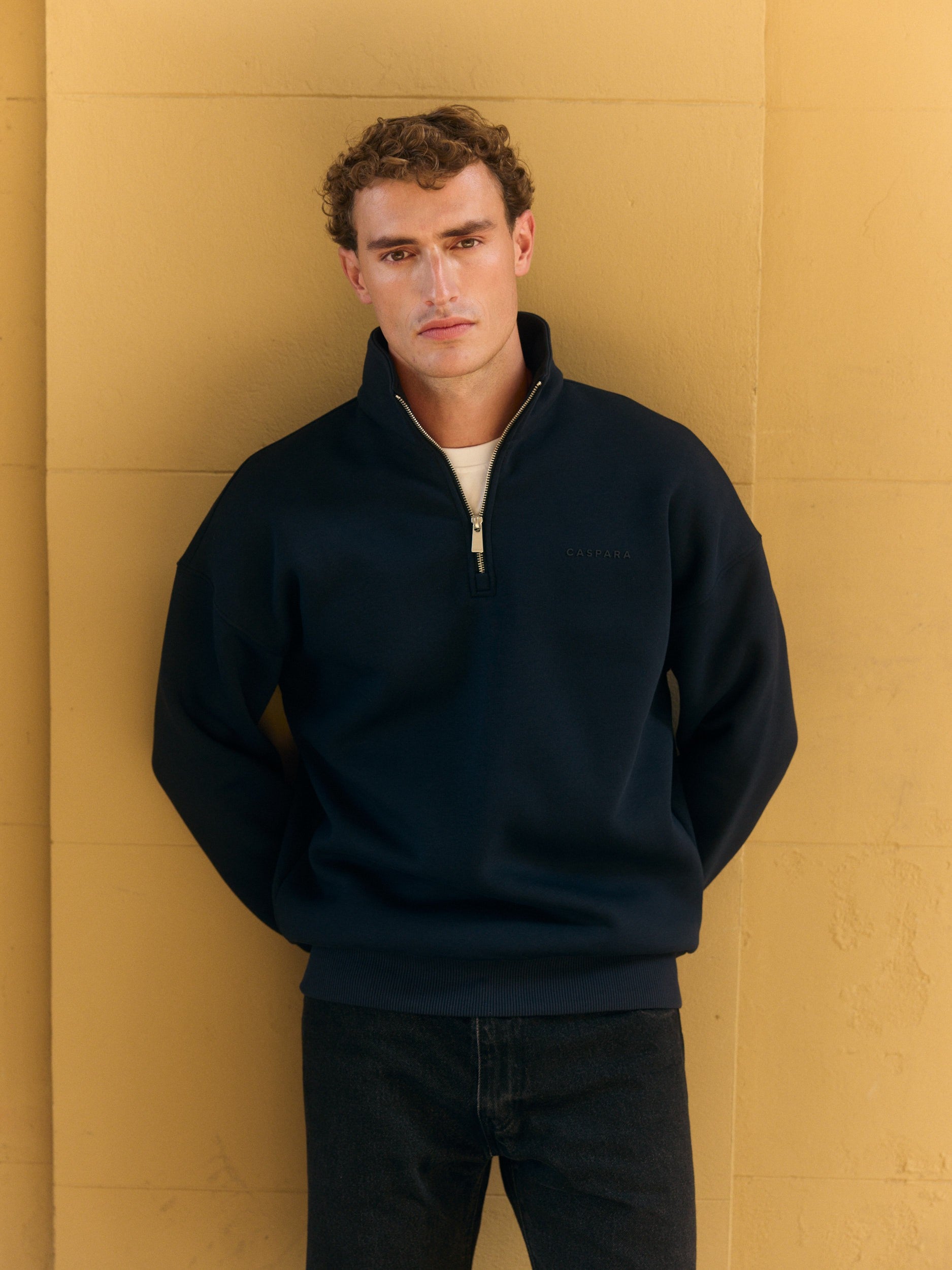 Man wearing a navy quarter-zip sweatshirt leaning against a yellow wall.