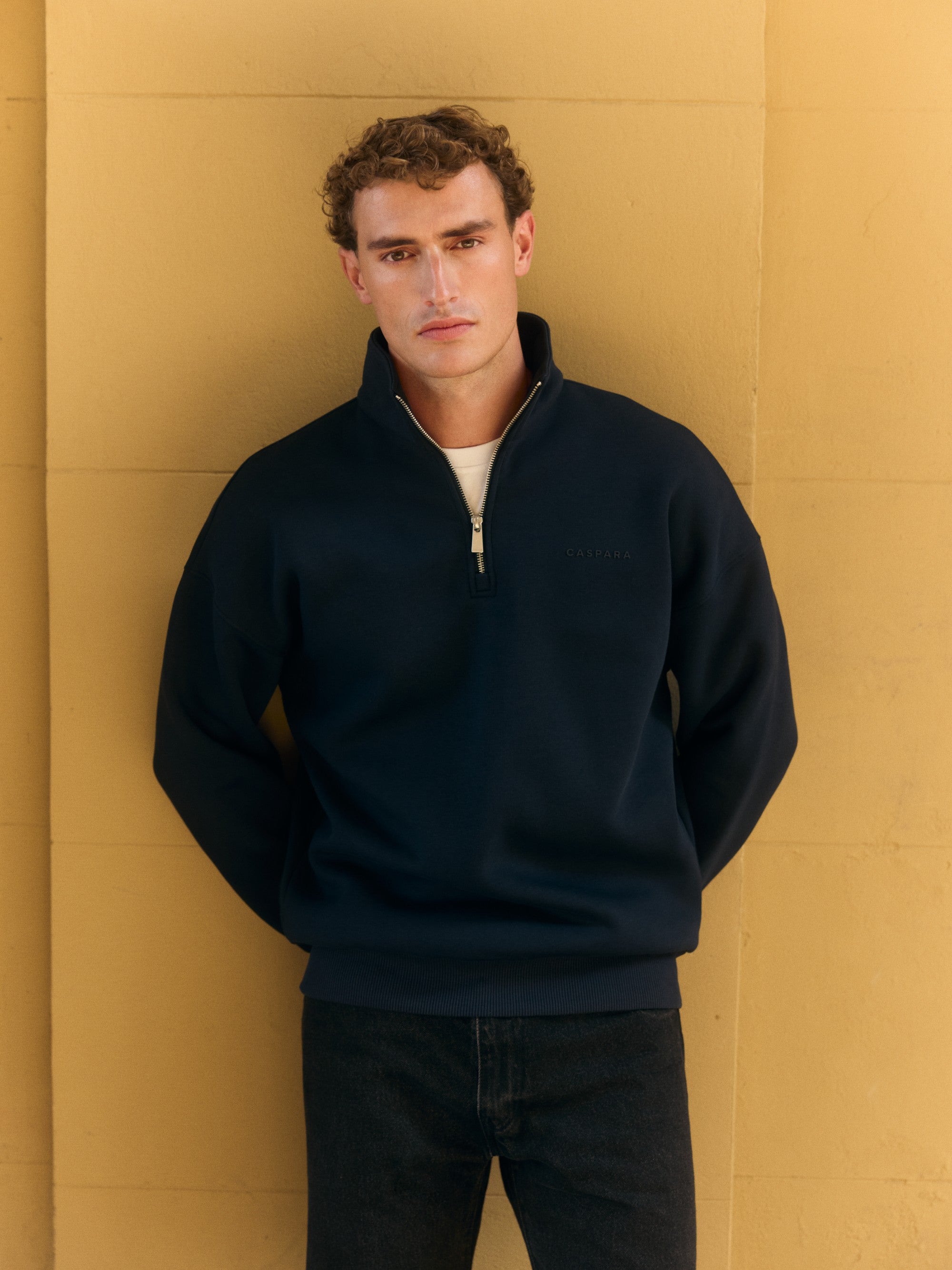 Man wearing a dark navy CASPARA quarter-zip sweatshirt against a yellow wall.