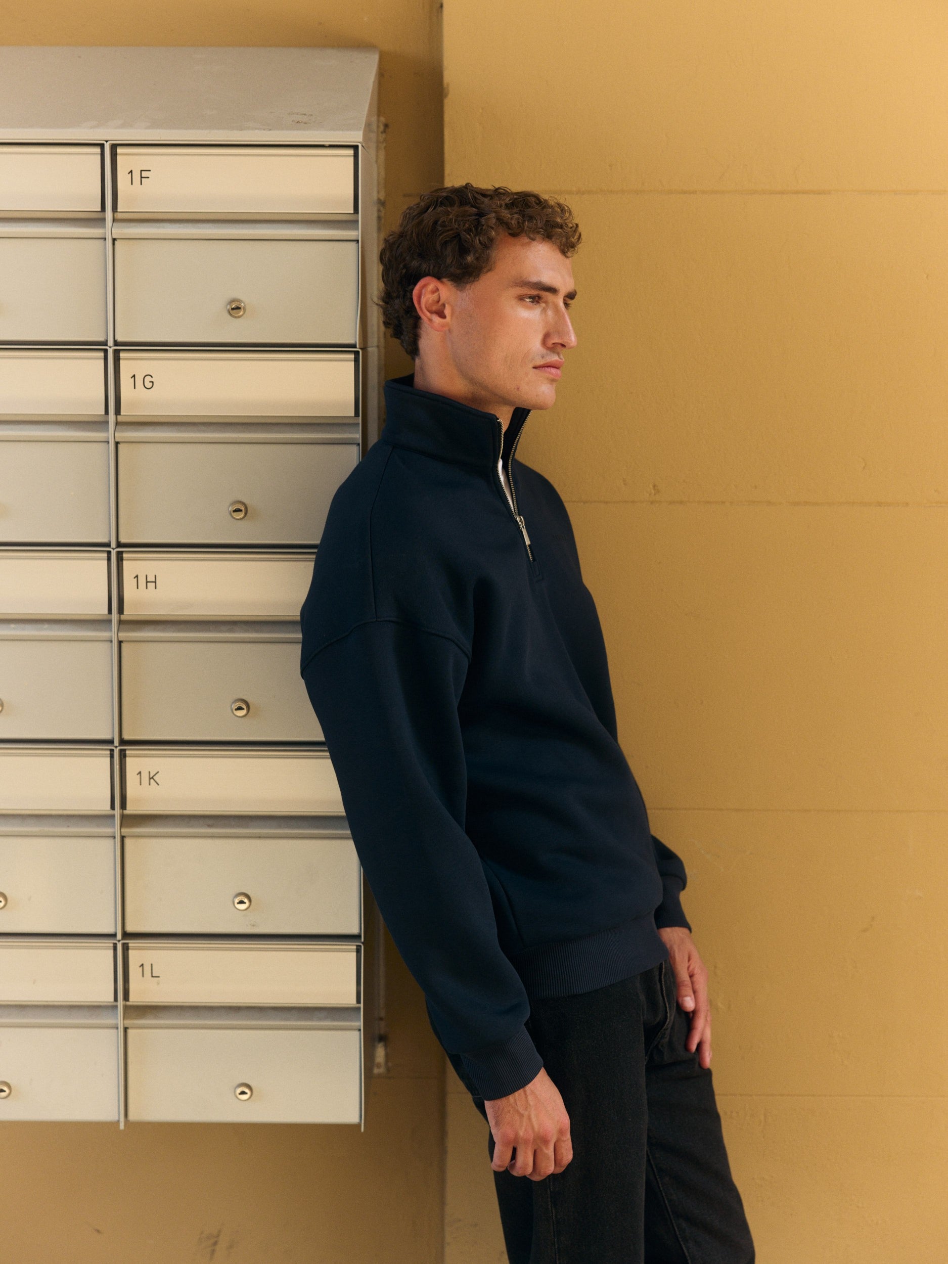 Man wearing a navy quarter-zip sweatshirt leaning against mailboxes by a yellow wall.