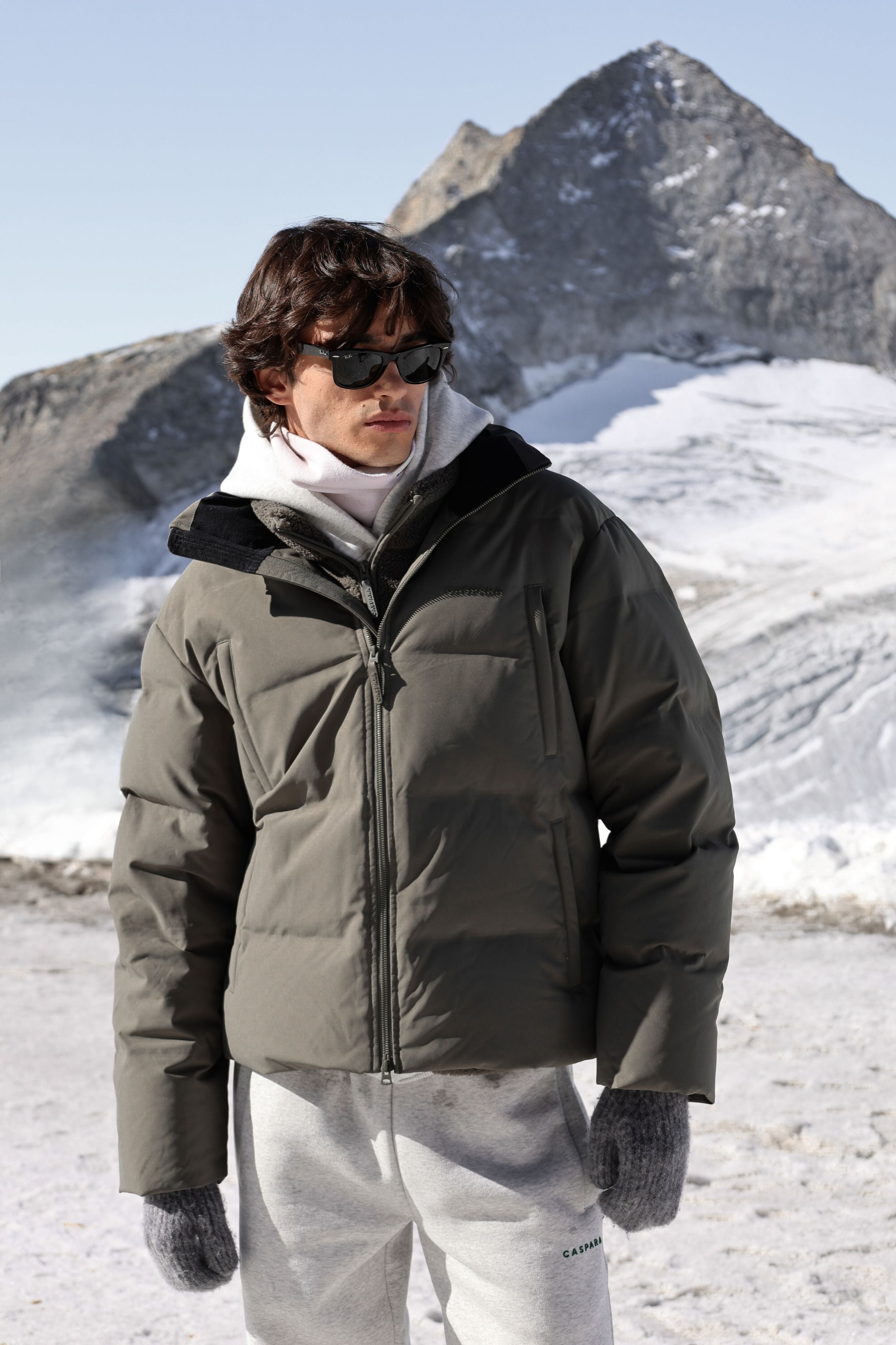 Man wearing a khaki CASPARA Boreal puffer jacket against a snowy mountain.