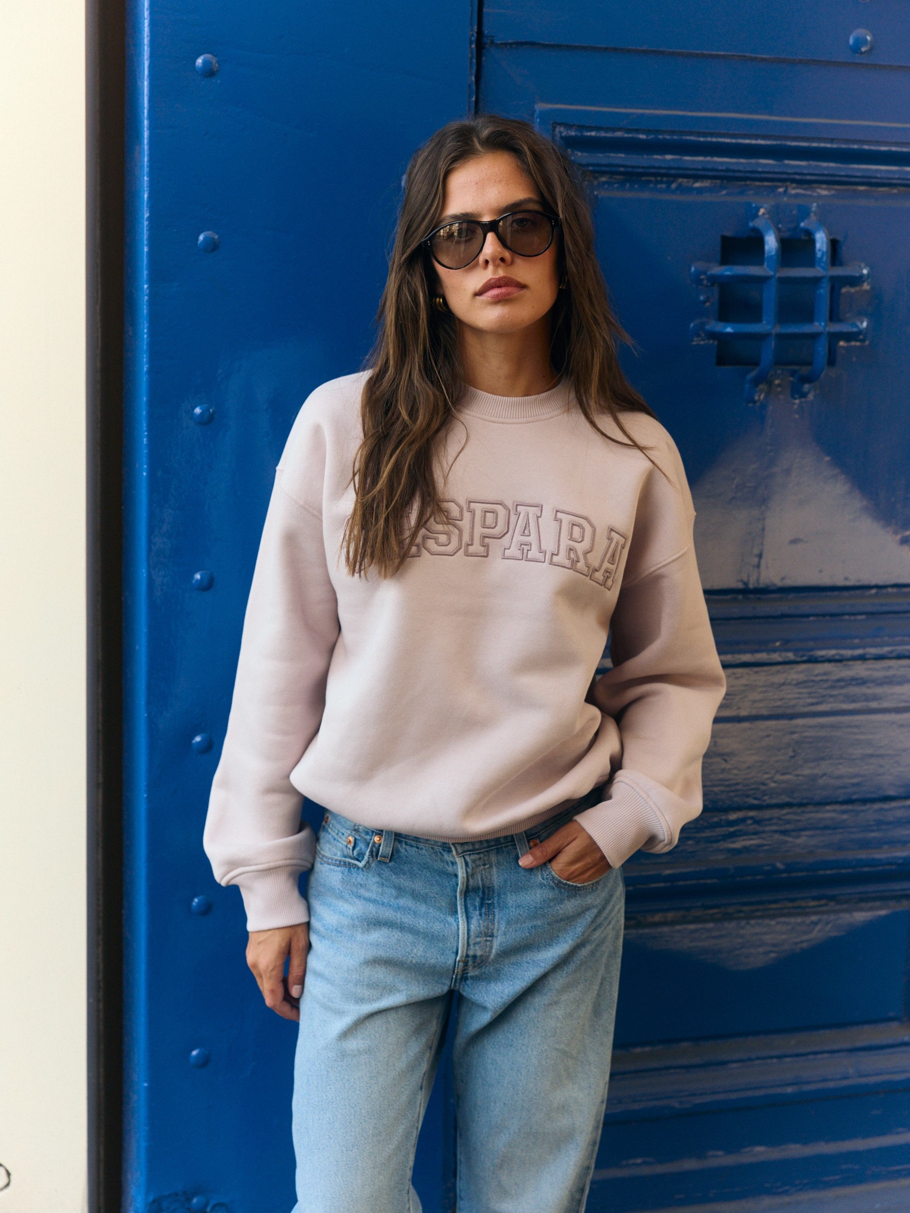 Woman wearing a dusty pink CASPARA crewneck sweatshirt and blue jeans against a blue door.