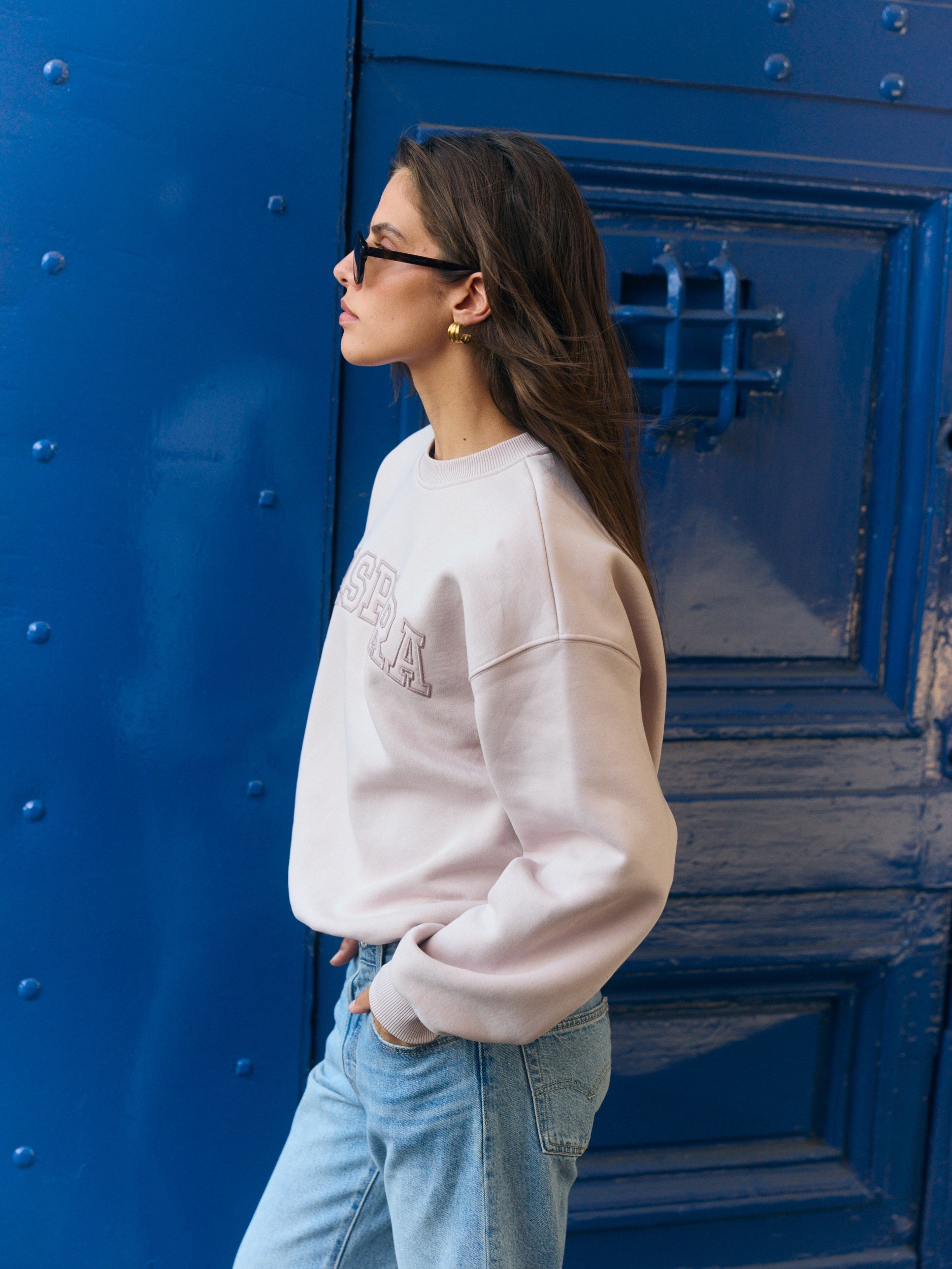 Woman wearing a dusty pink oversized crewneck sweatshirt and jeans in front of a blue door.