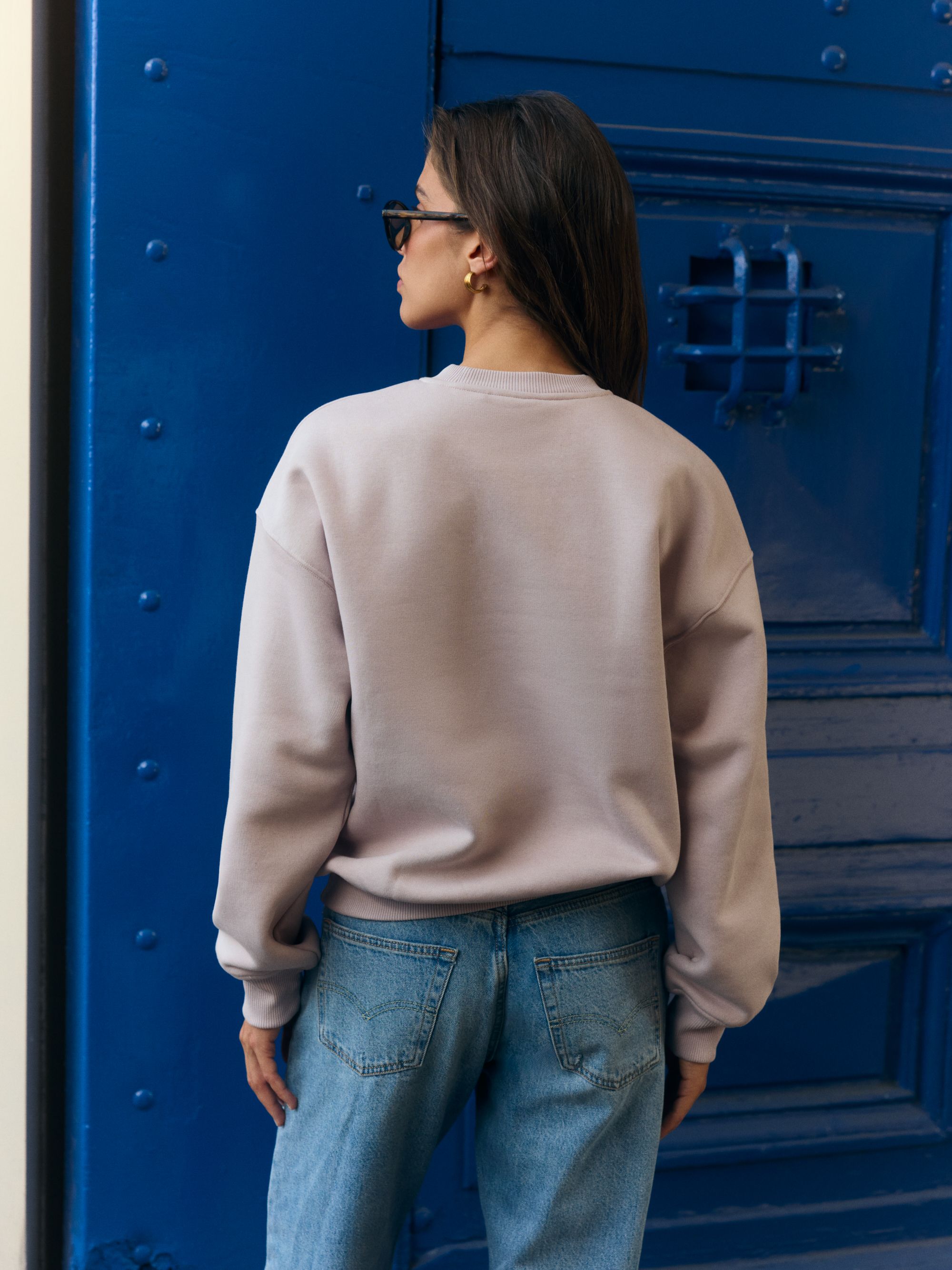 Back view of a woman wearing a dusty pink CASPARA crewneck sweatshirt in front of a blue door.
