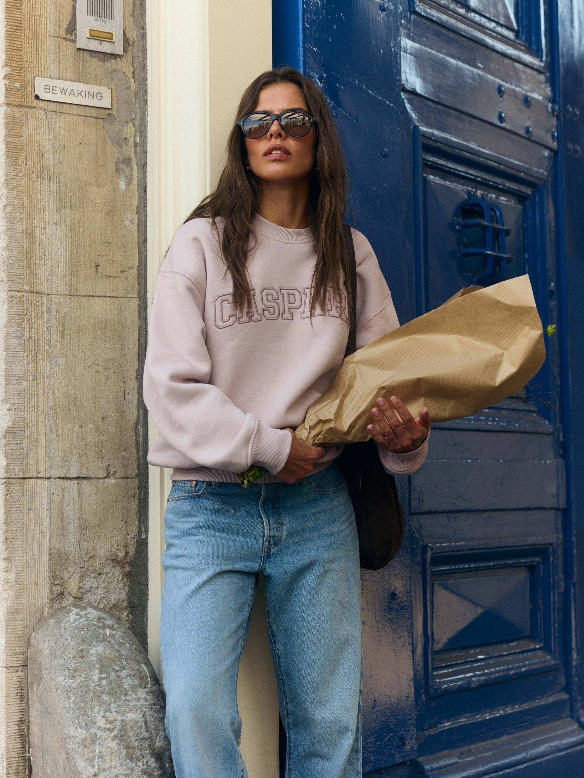 Woman wearing a dusty pink CASPARA crewneck sweatshirt and blue jeans holding flowers outside a blue door.