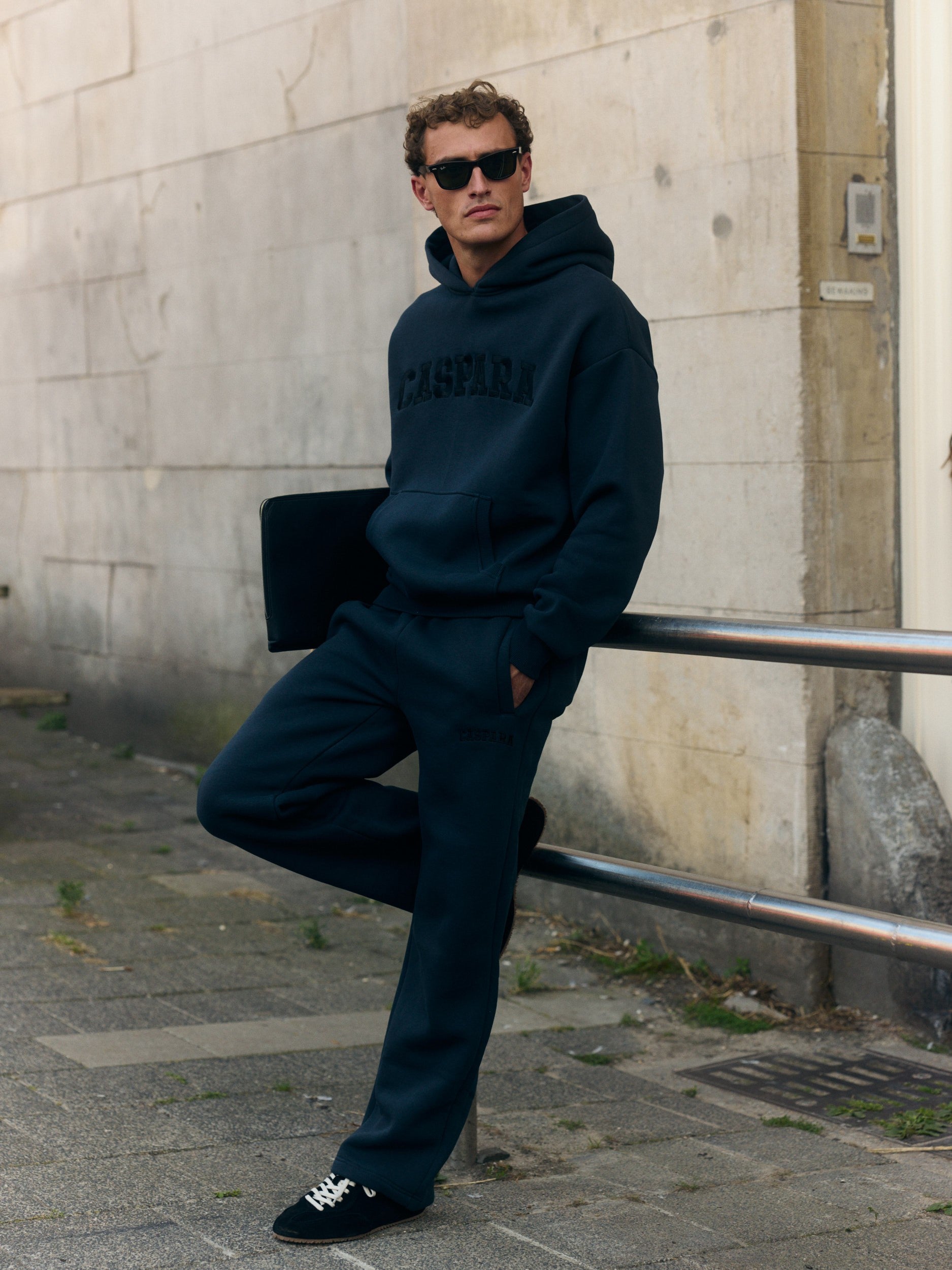 Man wearing a black CASPARA hoodie and sweatpants leaning against a metal railing on a city street.