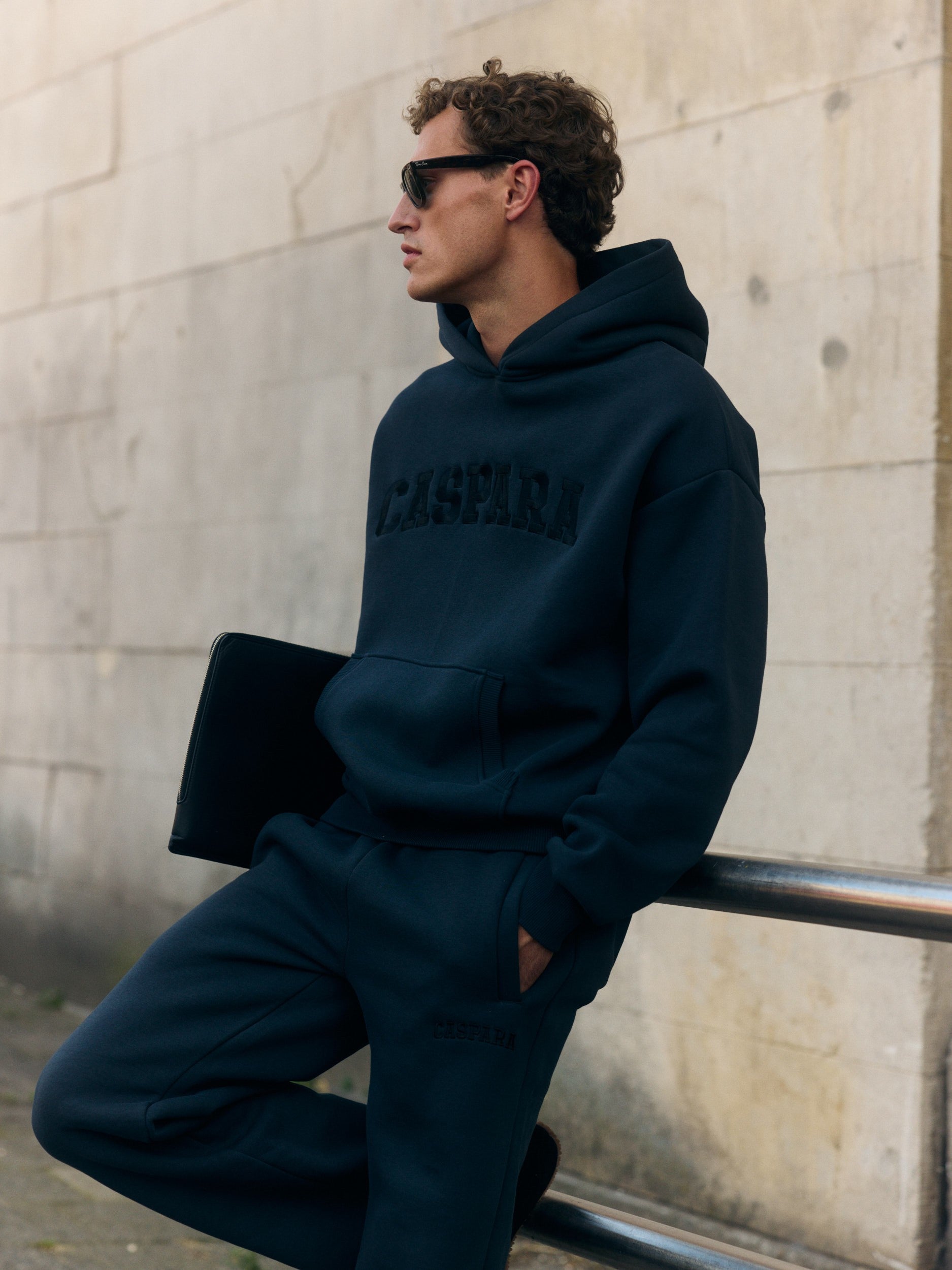 Man wearing a black CASPARA hoodie and matching sweatpants leaning against a railing.