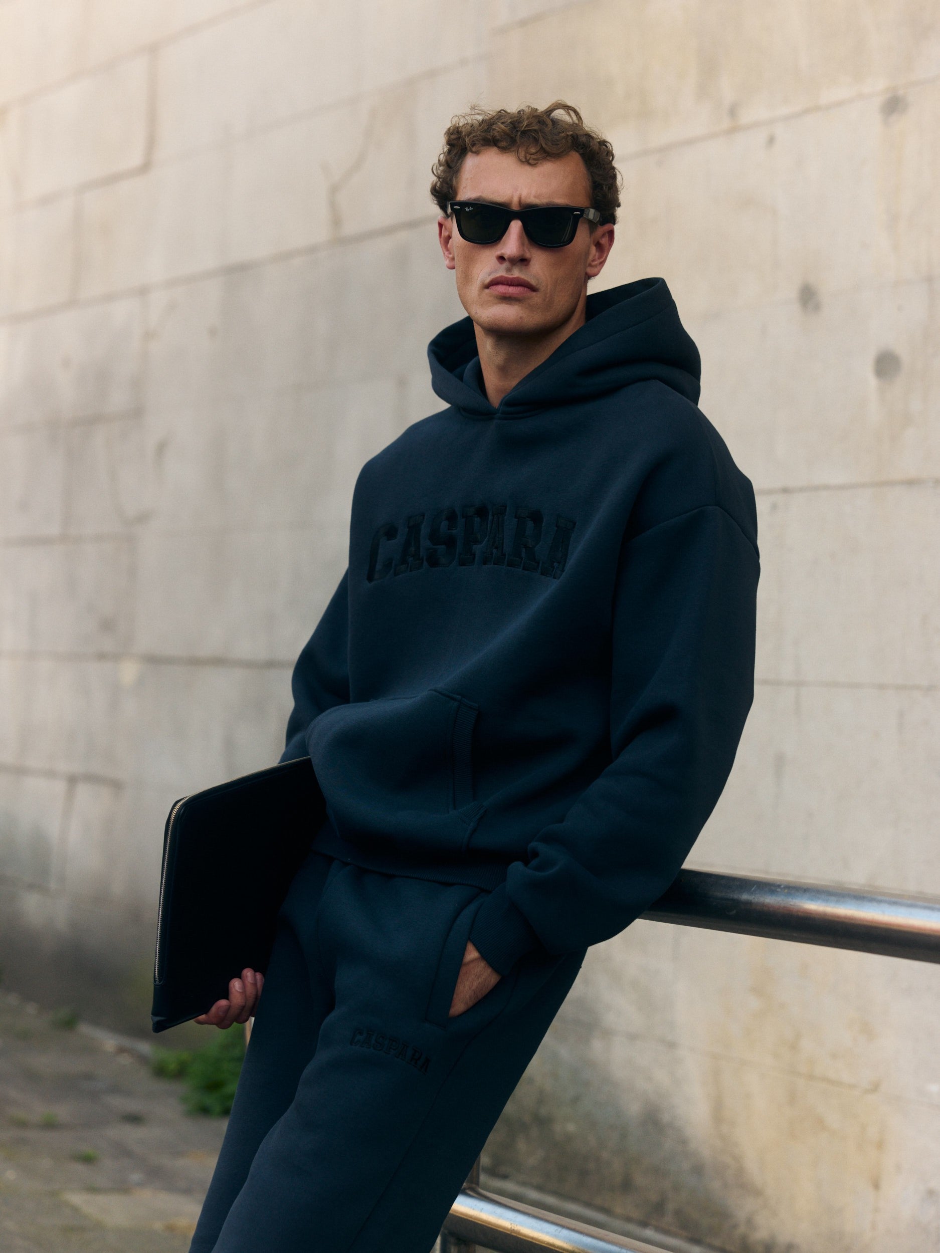 Man wearing a black CASPARA hoodie and matching sweatpants leaning against a railing.