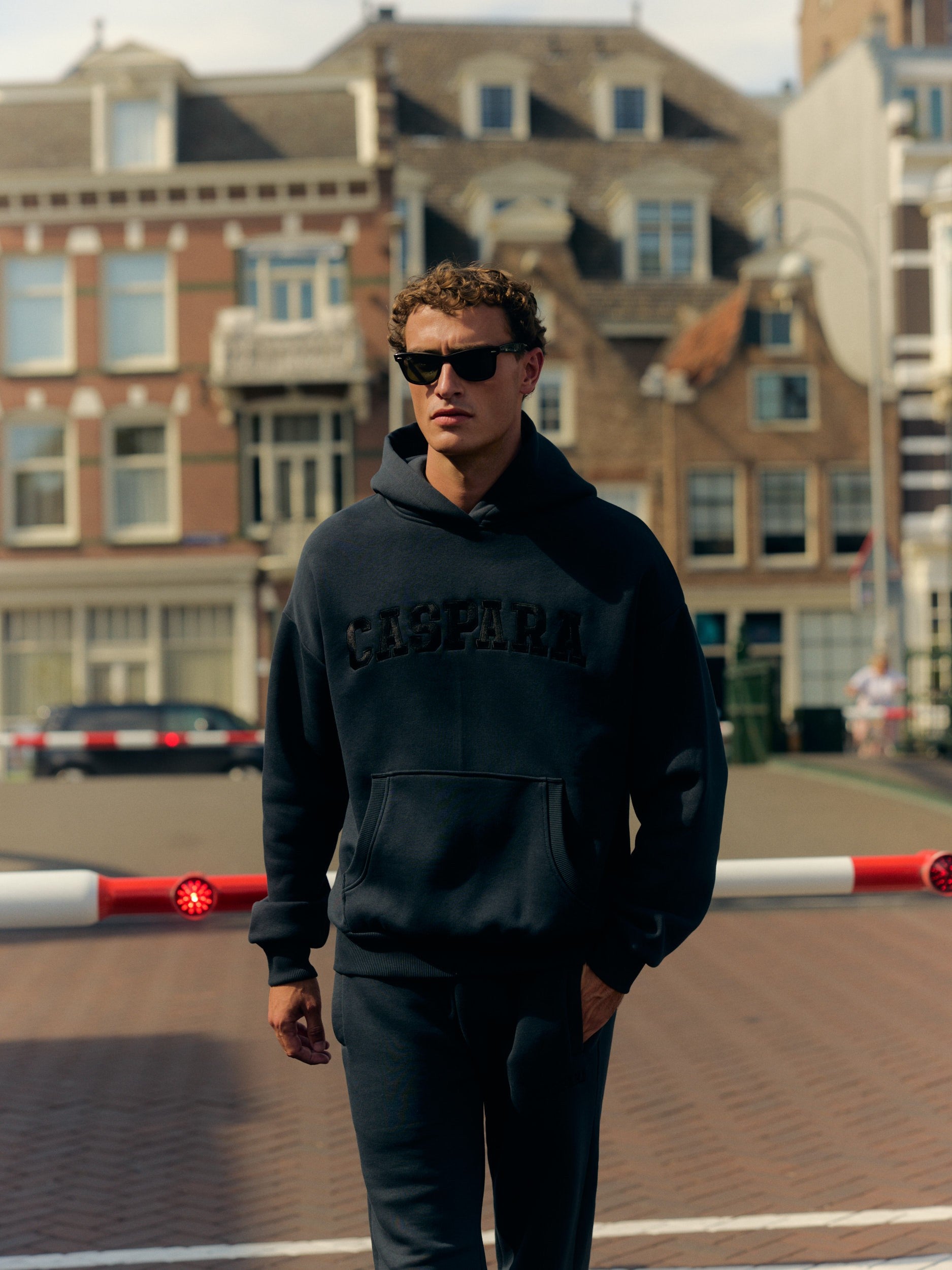 Man wearing a black CASPARA logo hoodie and sunglasses walking on a city street.