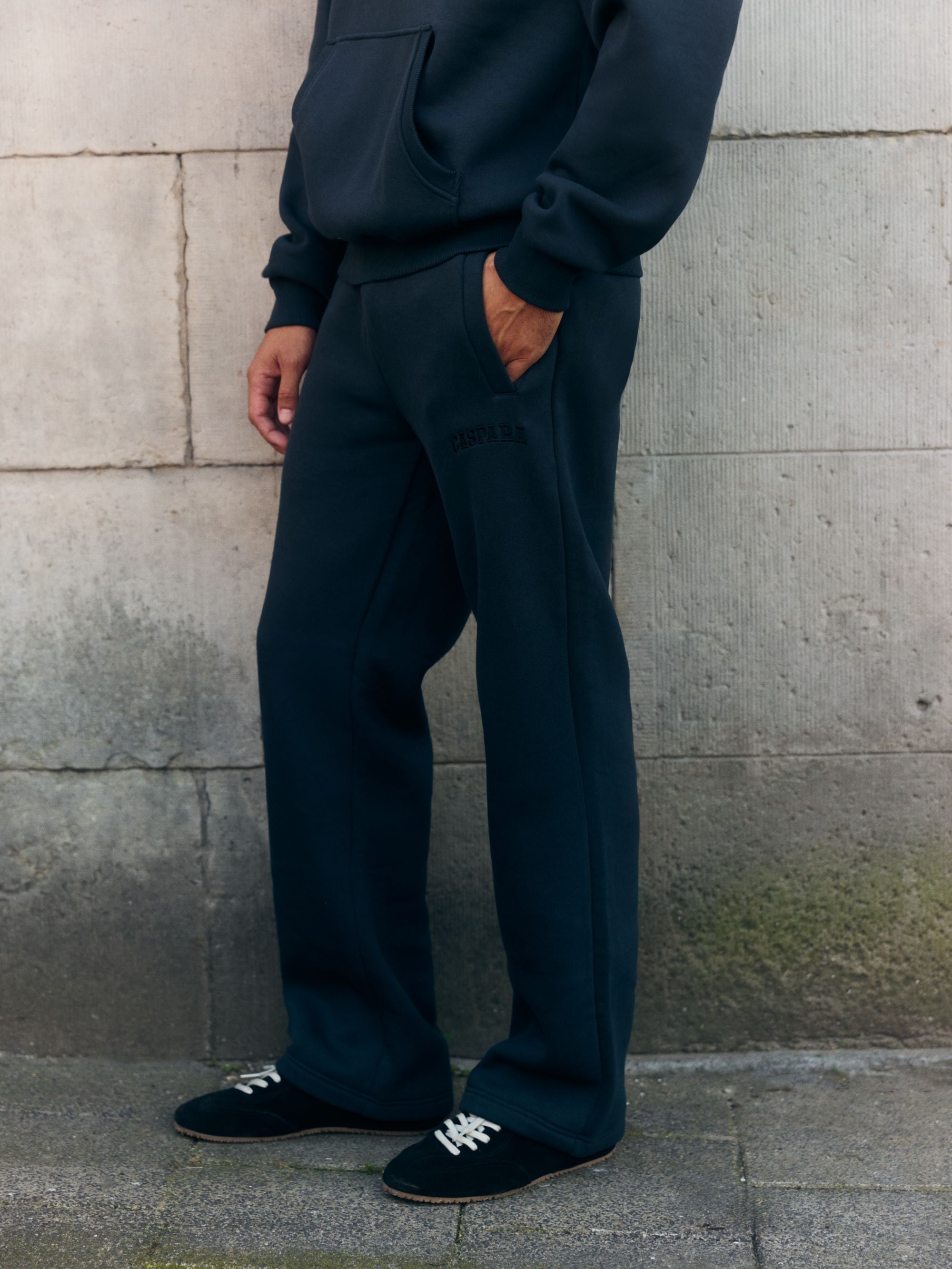 Man wearing dark navy CASPARA straight-leg sweatpants and a matching hoodie against a stone wall.