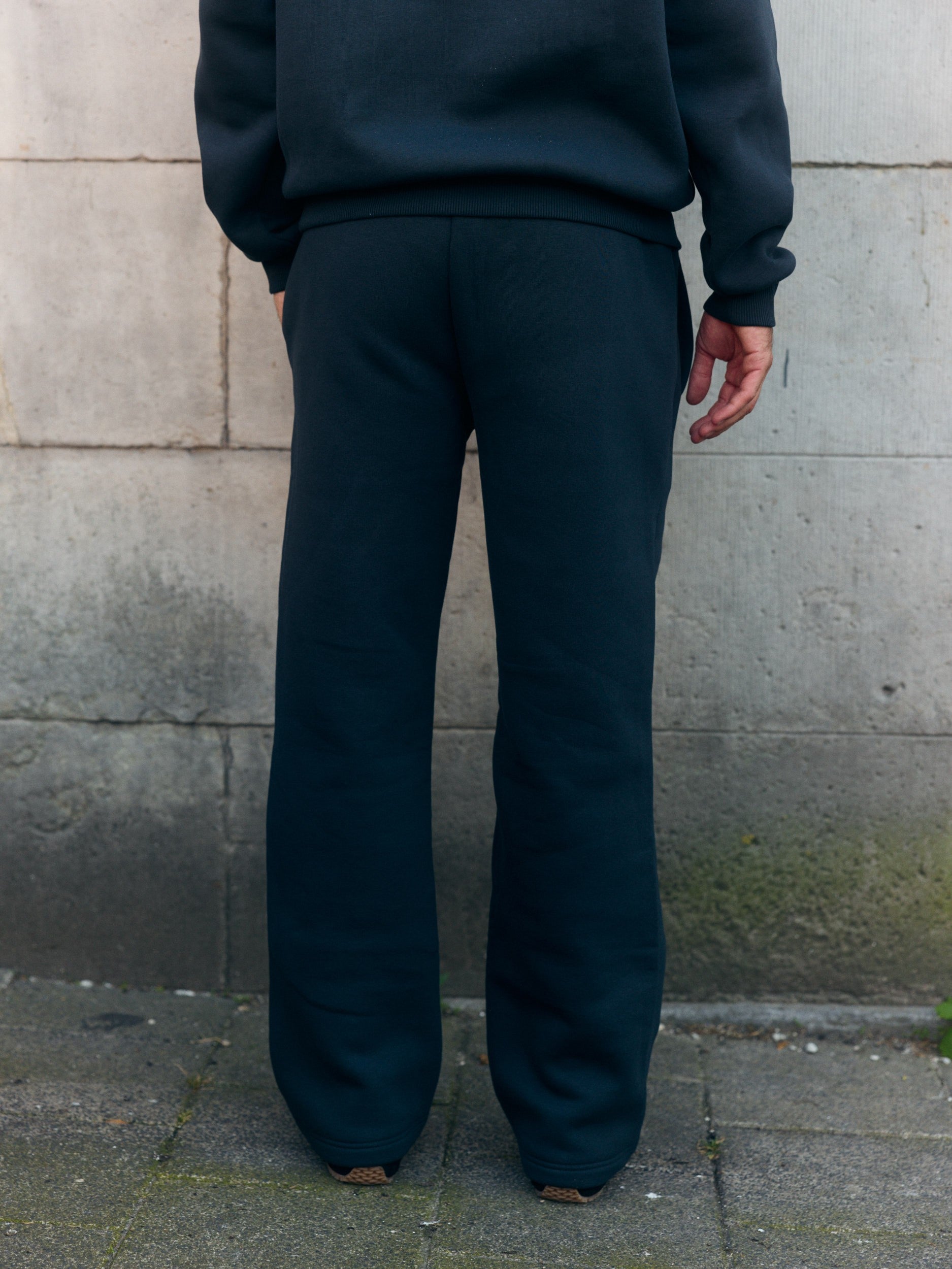 Rear view of a man wearing black straight-leg sweatpants against an outdoor stone wall.