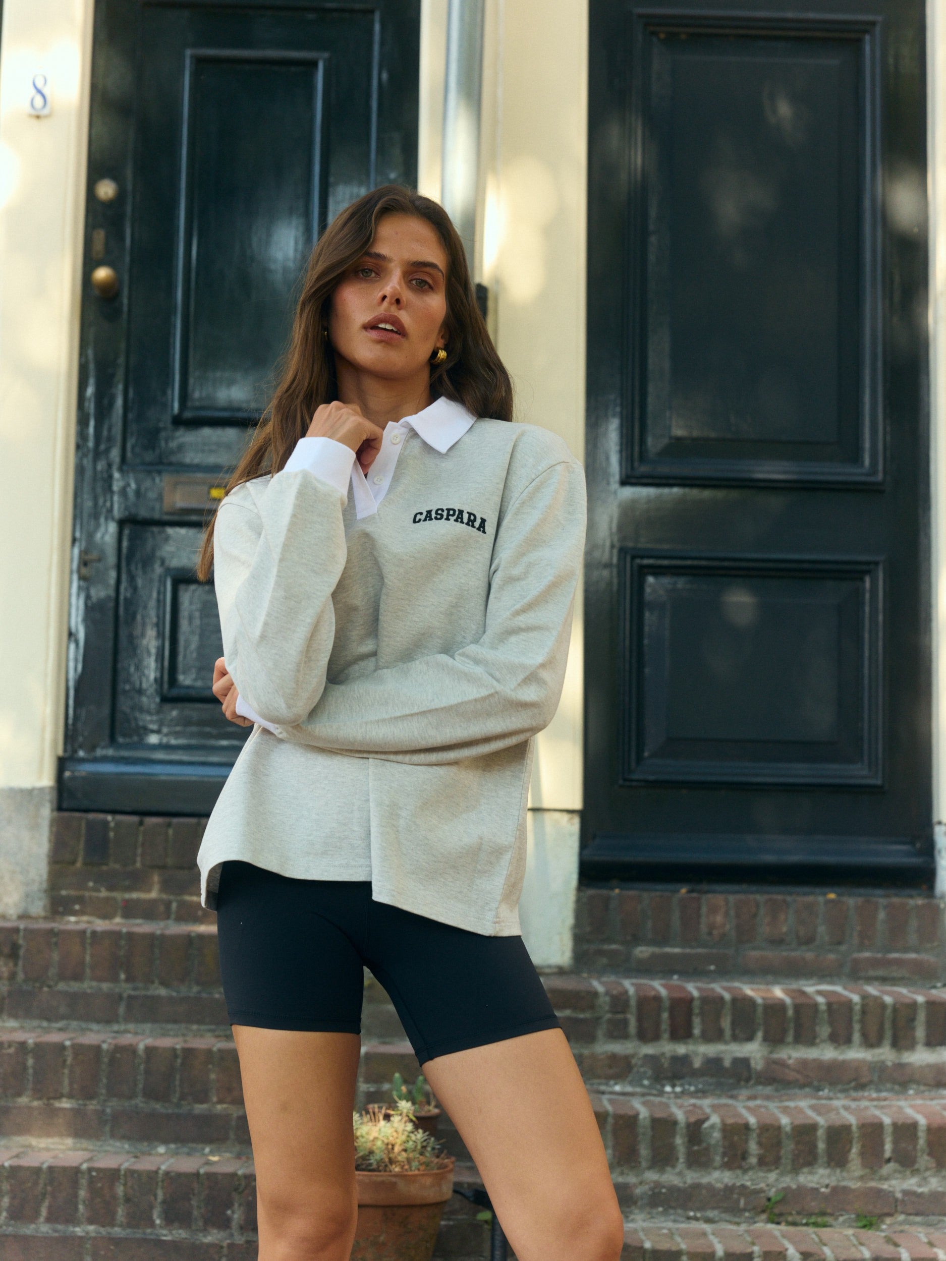 Woman wearing a light grey long-sleeve rugby shirt with a white collar outside a dark doorway.