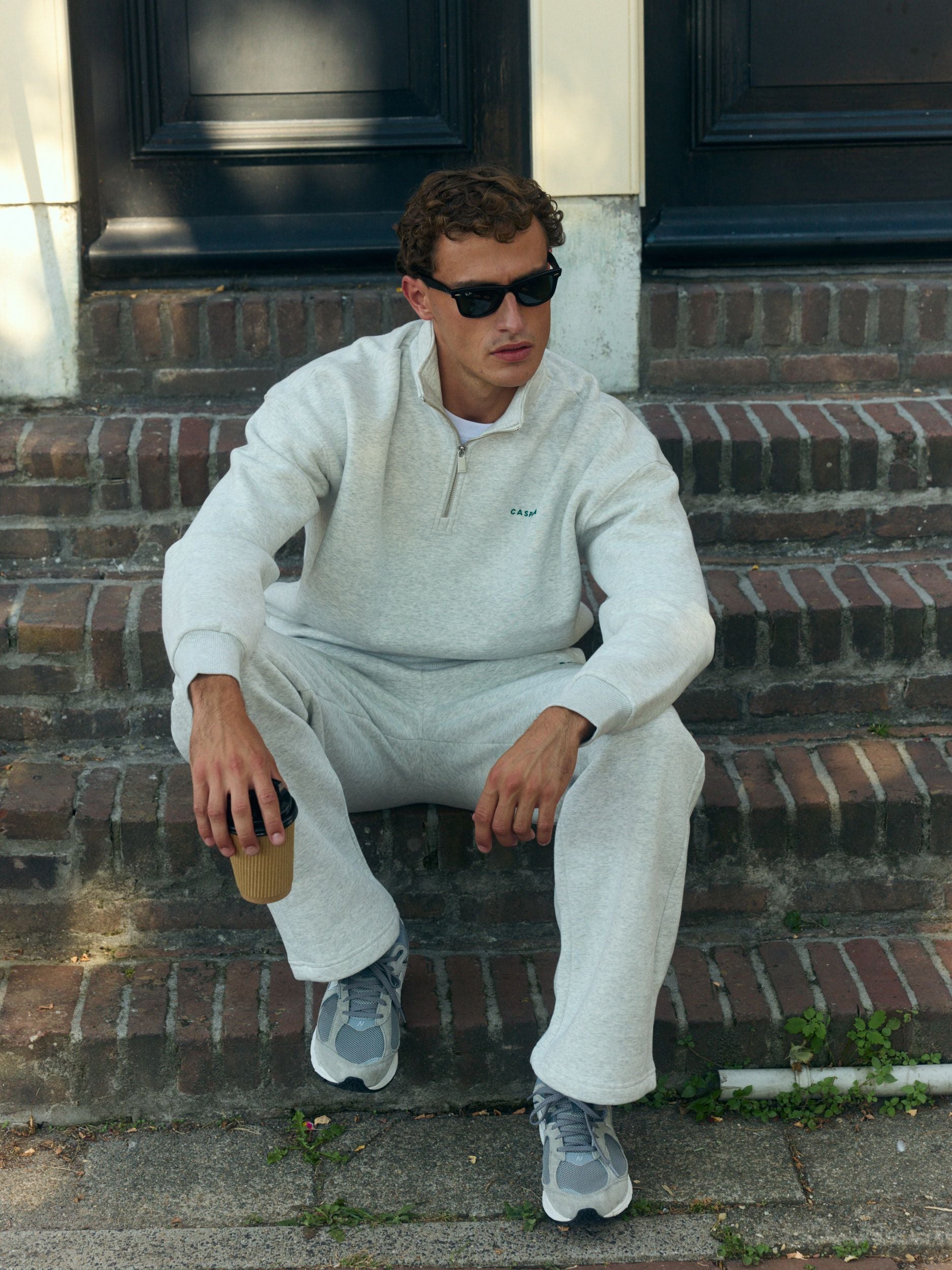 Man wearing a grey melange quarter-zip sweatshirt sitting on brick steps with a coffee cup.