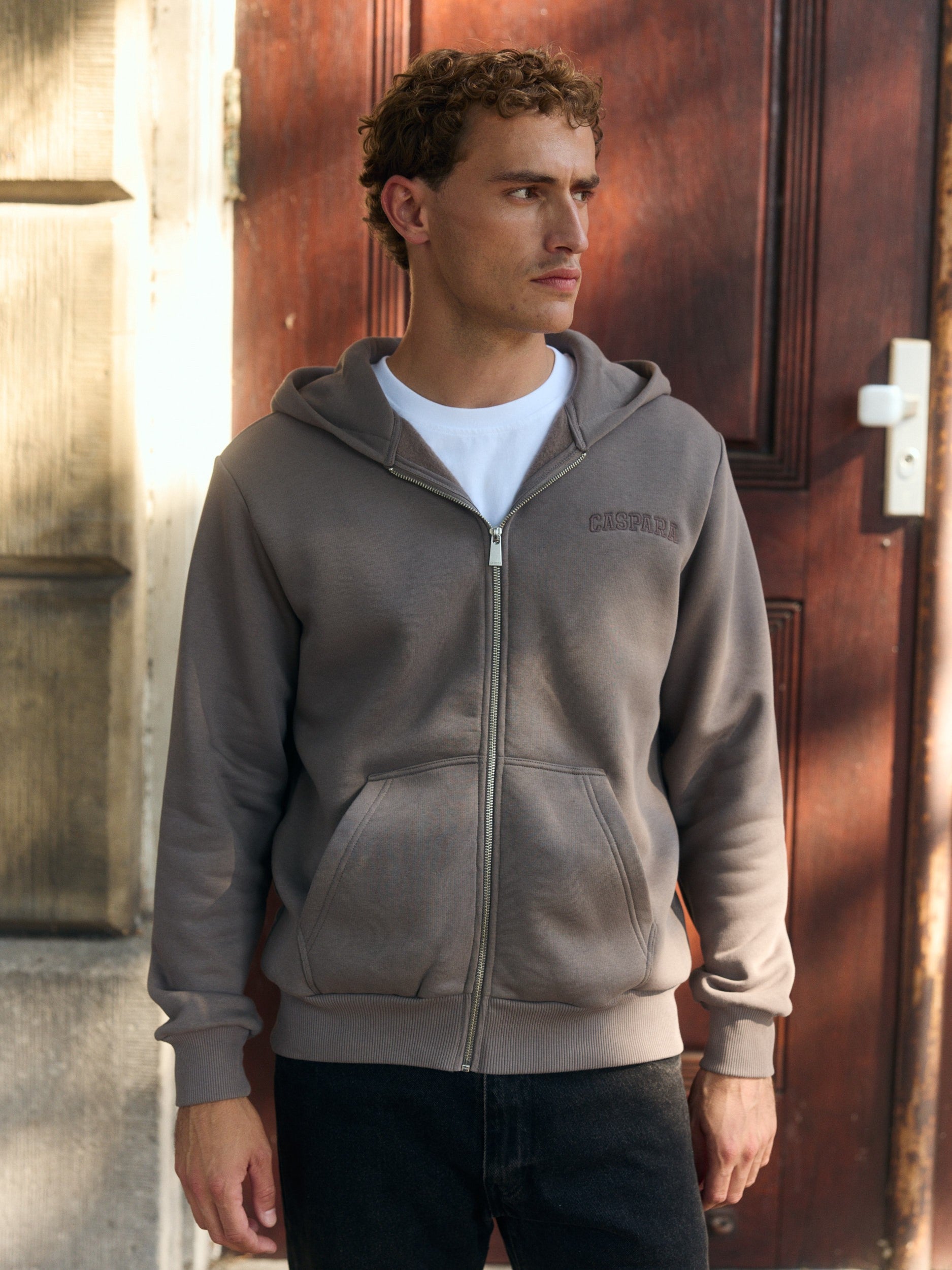 Man wearing a walnut brown zip hoodie over a white t-shirt standing in front of a wooden door.
