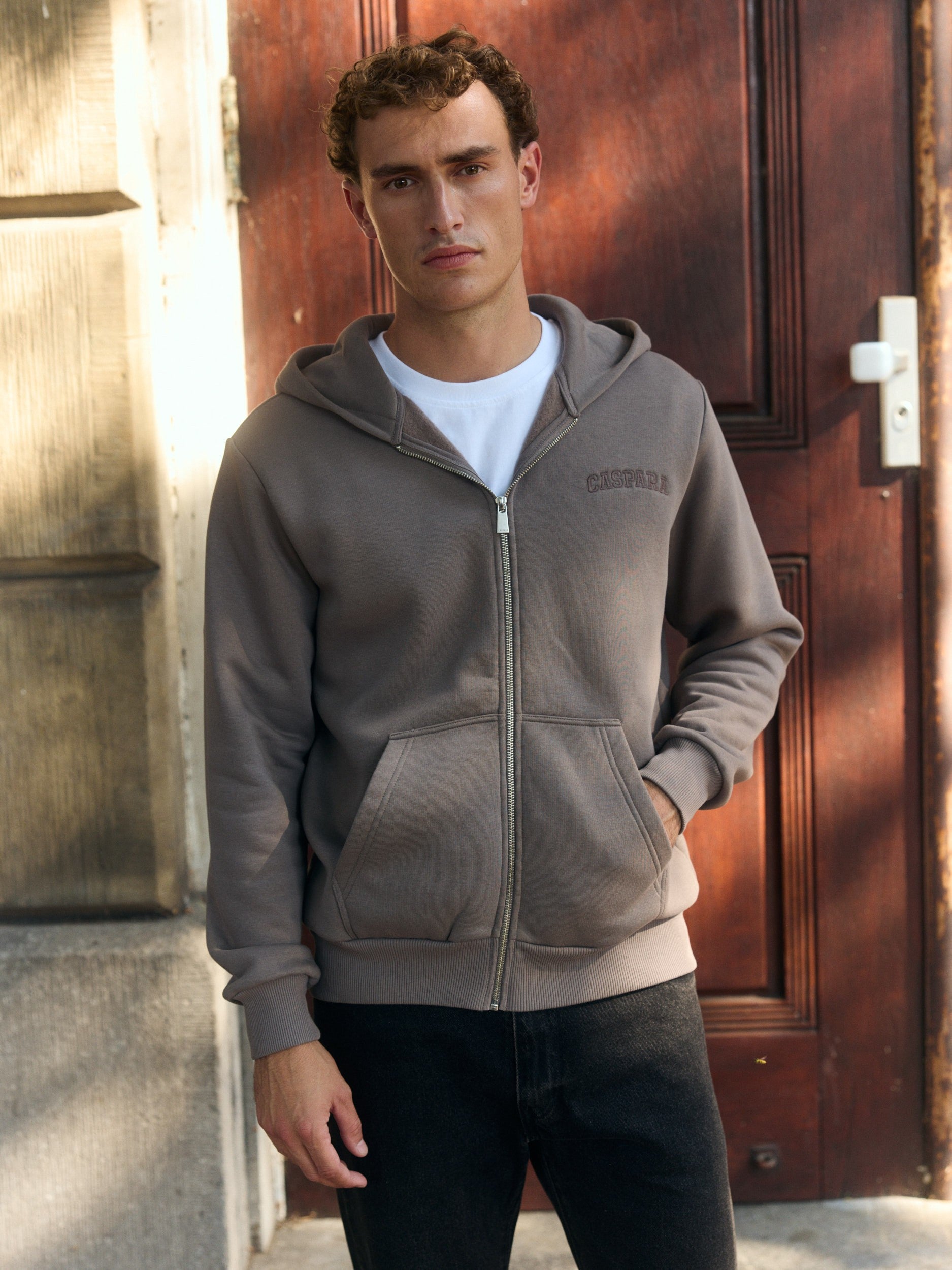 Man wearing a walnut zip hoodie over a white t-shirt in front of a wooden door.