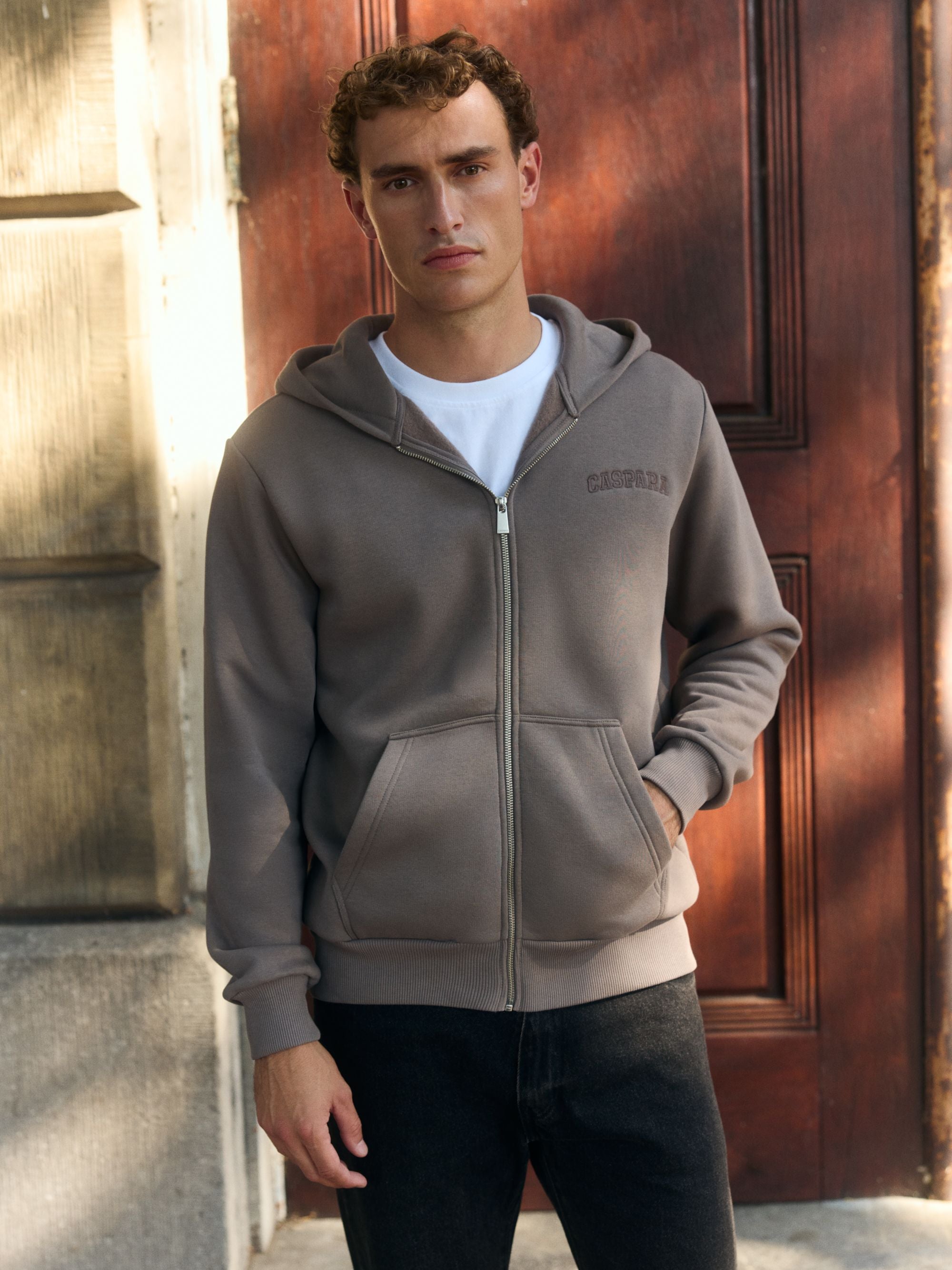 Man wearing a walnut CASPARA zip hoodie over a white t-shirt in front of a wooden door.