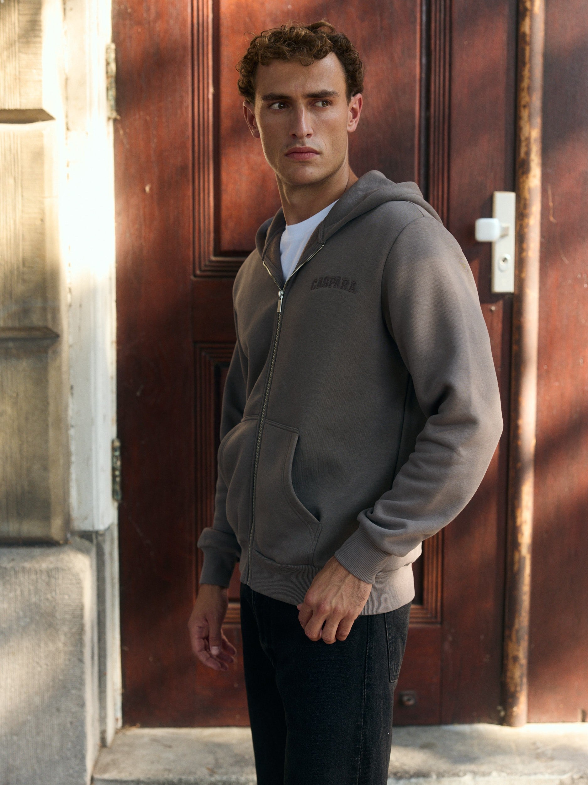 Man wearing a walnut brown zip hoodie standing in front of a wooden door.
