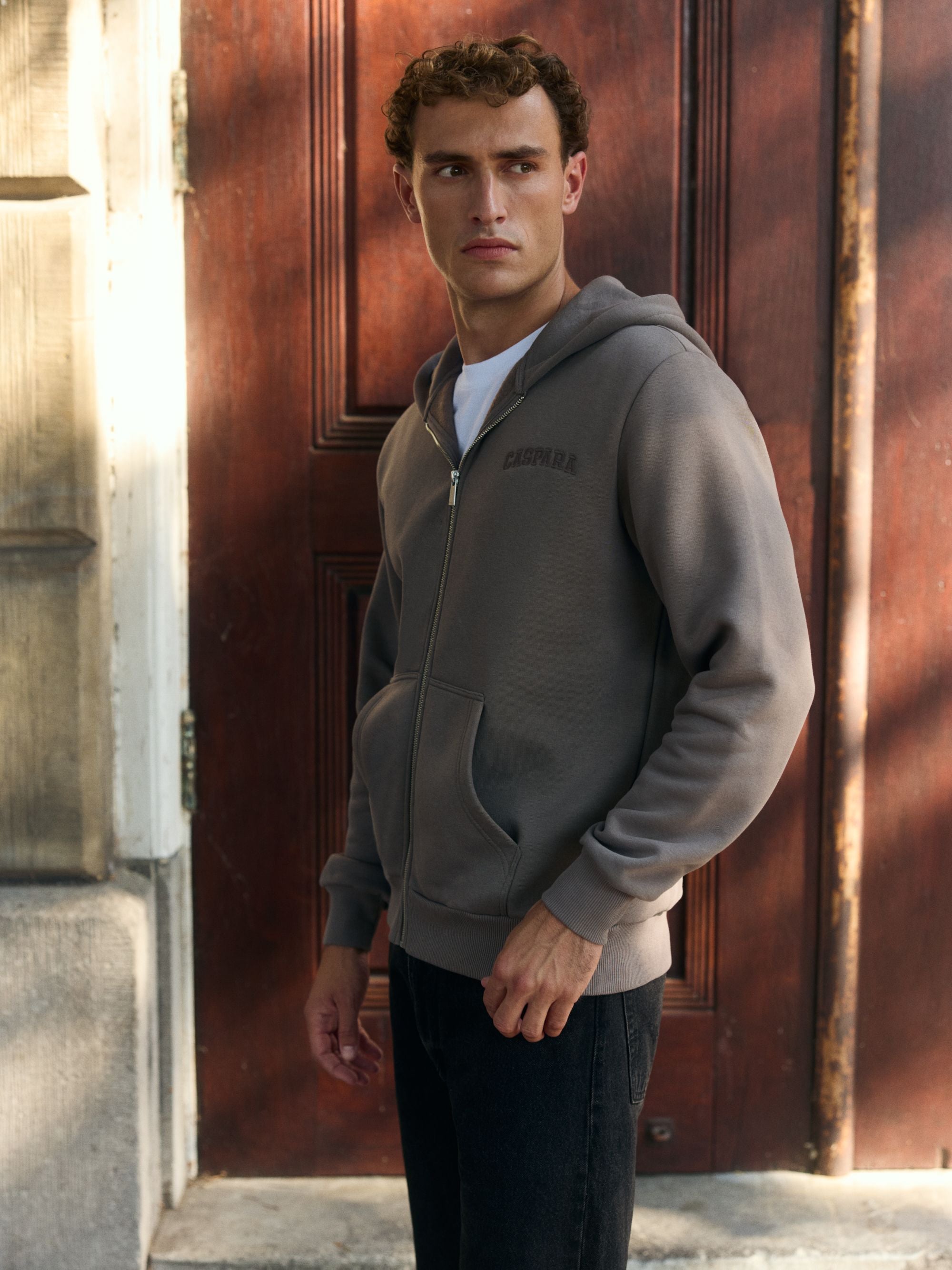 Man wearing a walnut CASPARA zip hoodie standing in front of a wooden door.