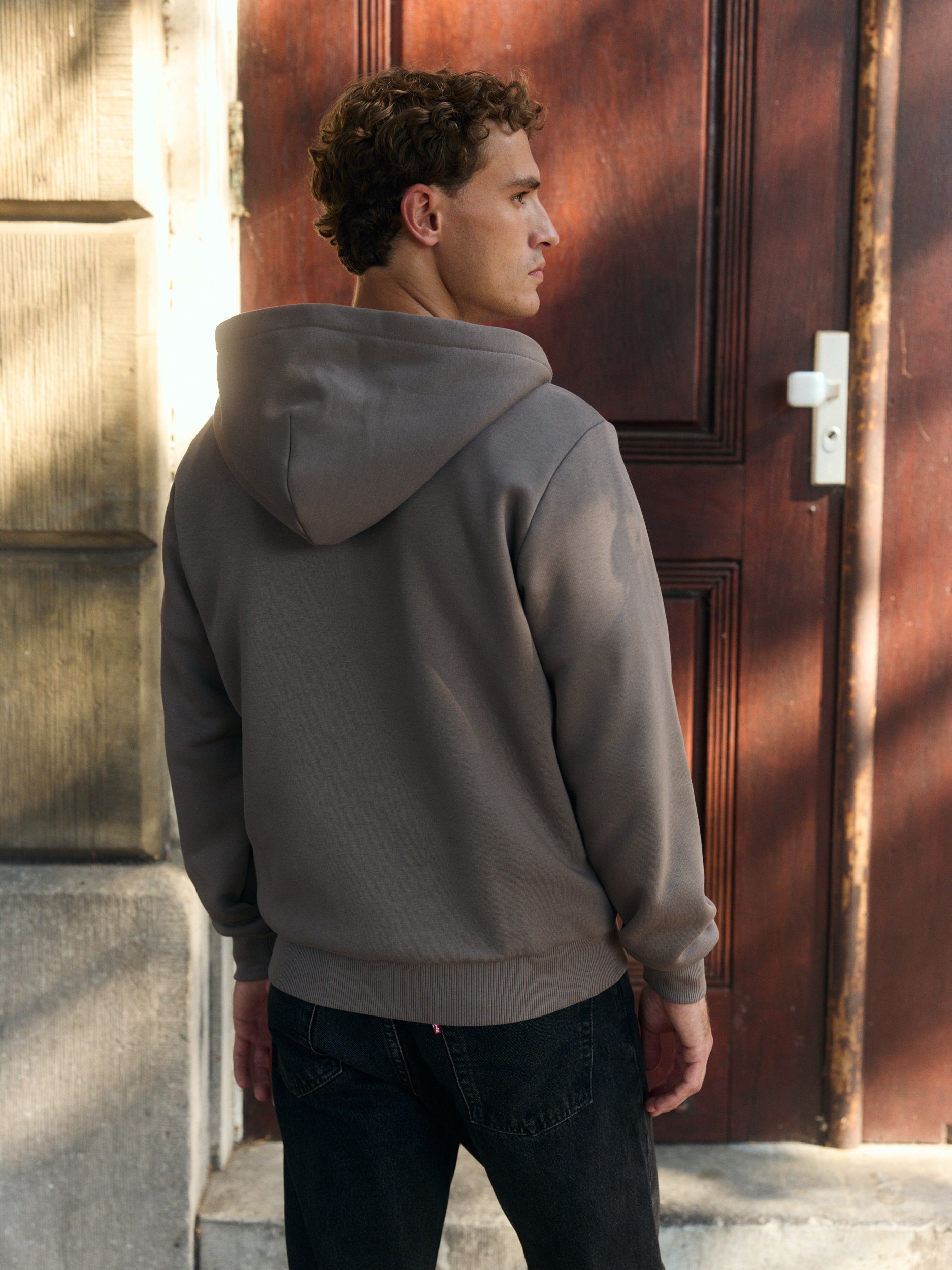 Man wearing a walnut zip hoodie seen from behind in front of a wooden door.