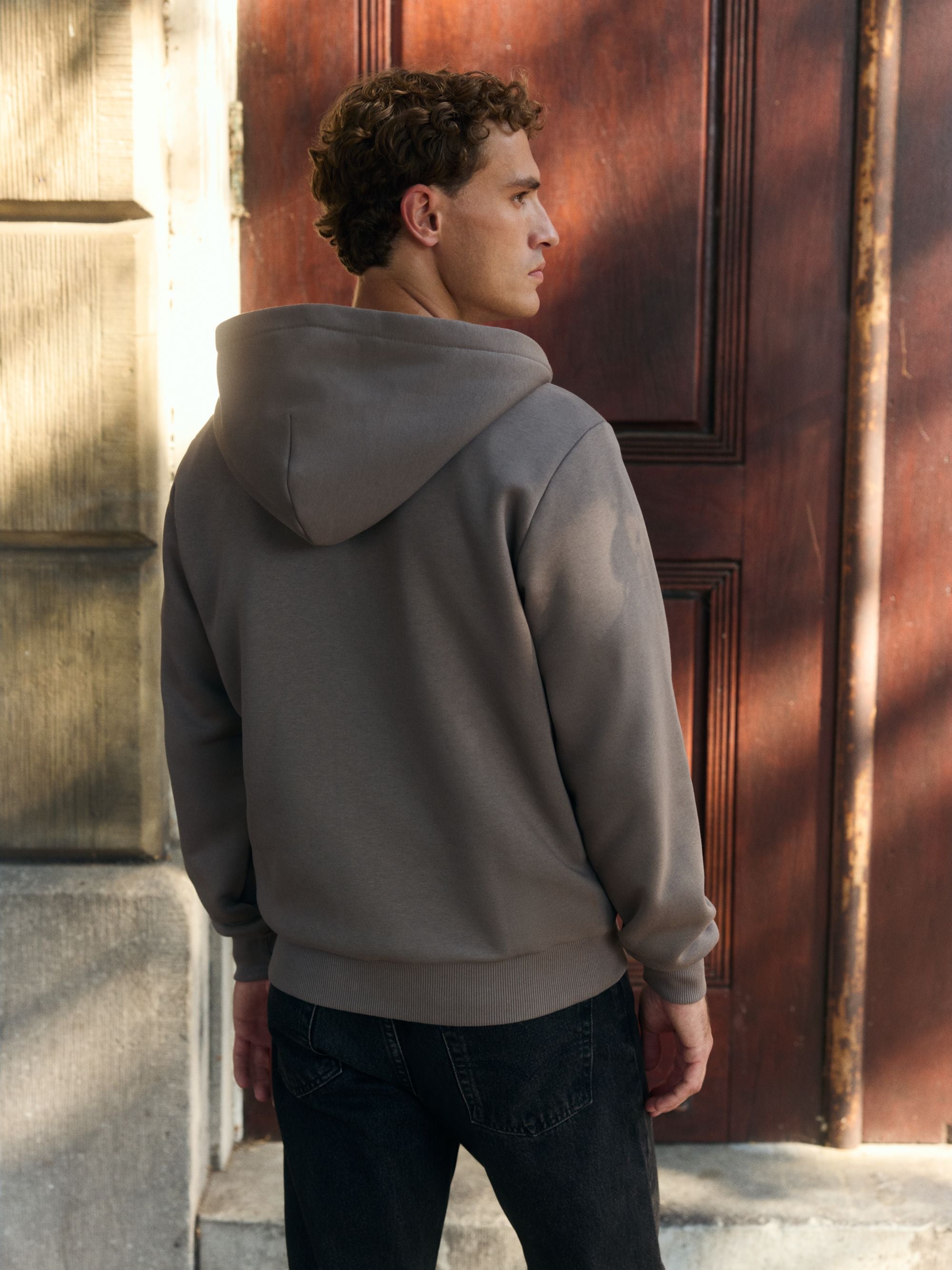 Back view of a man wearing a walnut CASPARA hoodie standing in front of a wooden door.