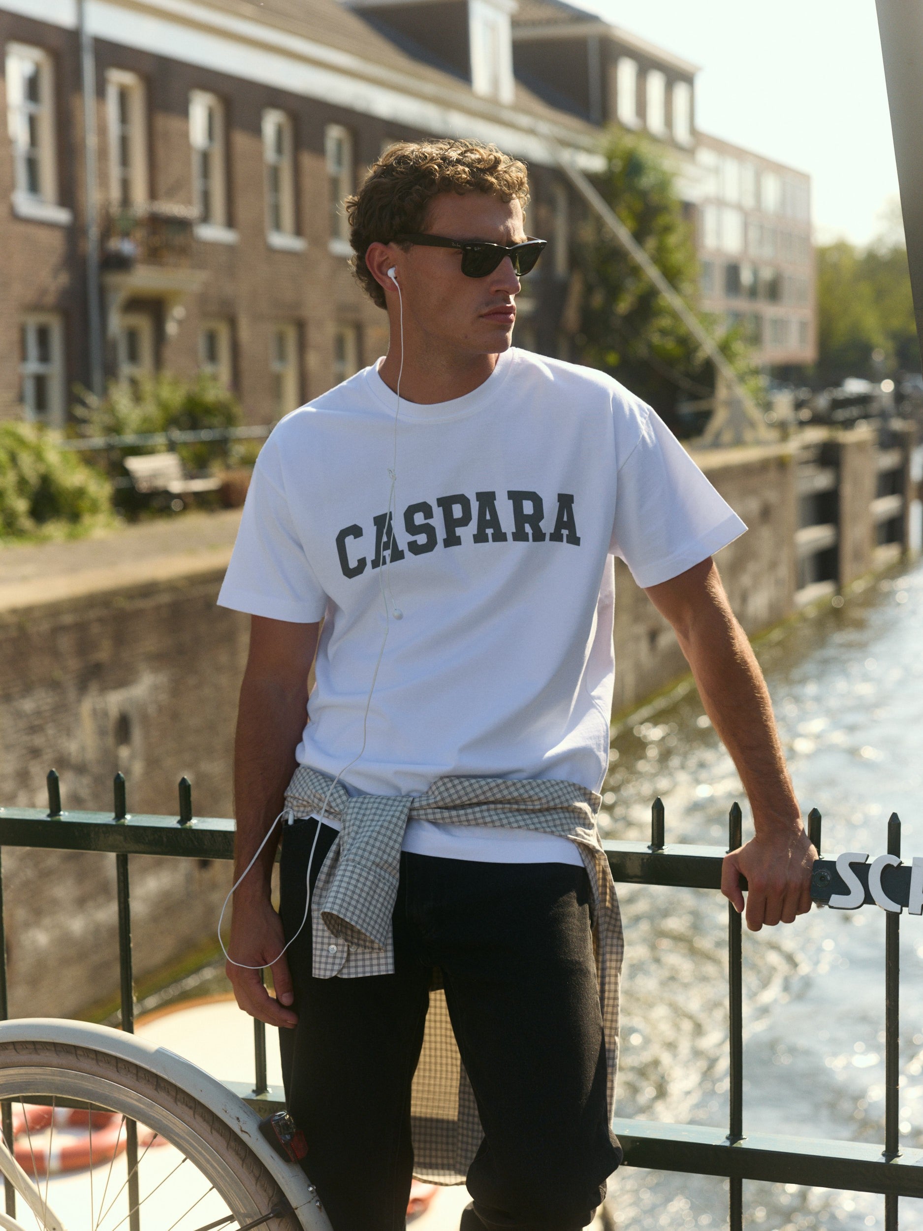 Man wearing a white CASPARA logo t-shirt and sunglasses leaning against a canal railing.