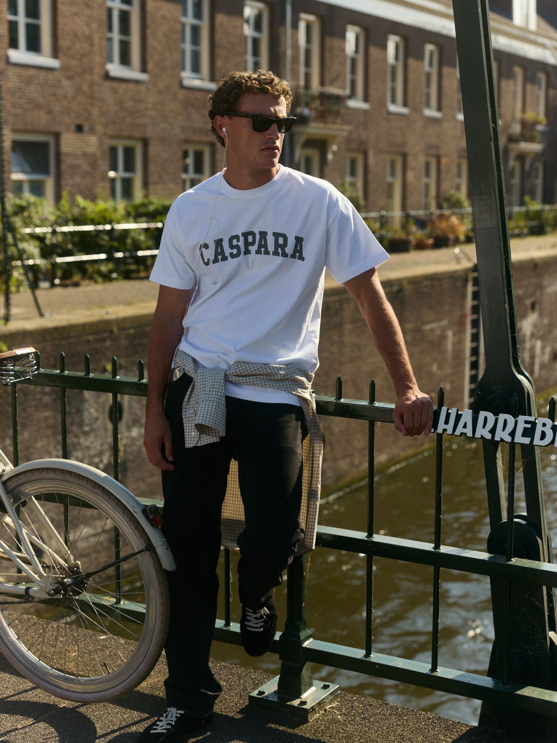Man wearing a white CASPARA logo t-shirt leaning against a canal bridge railing.