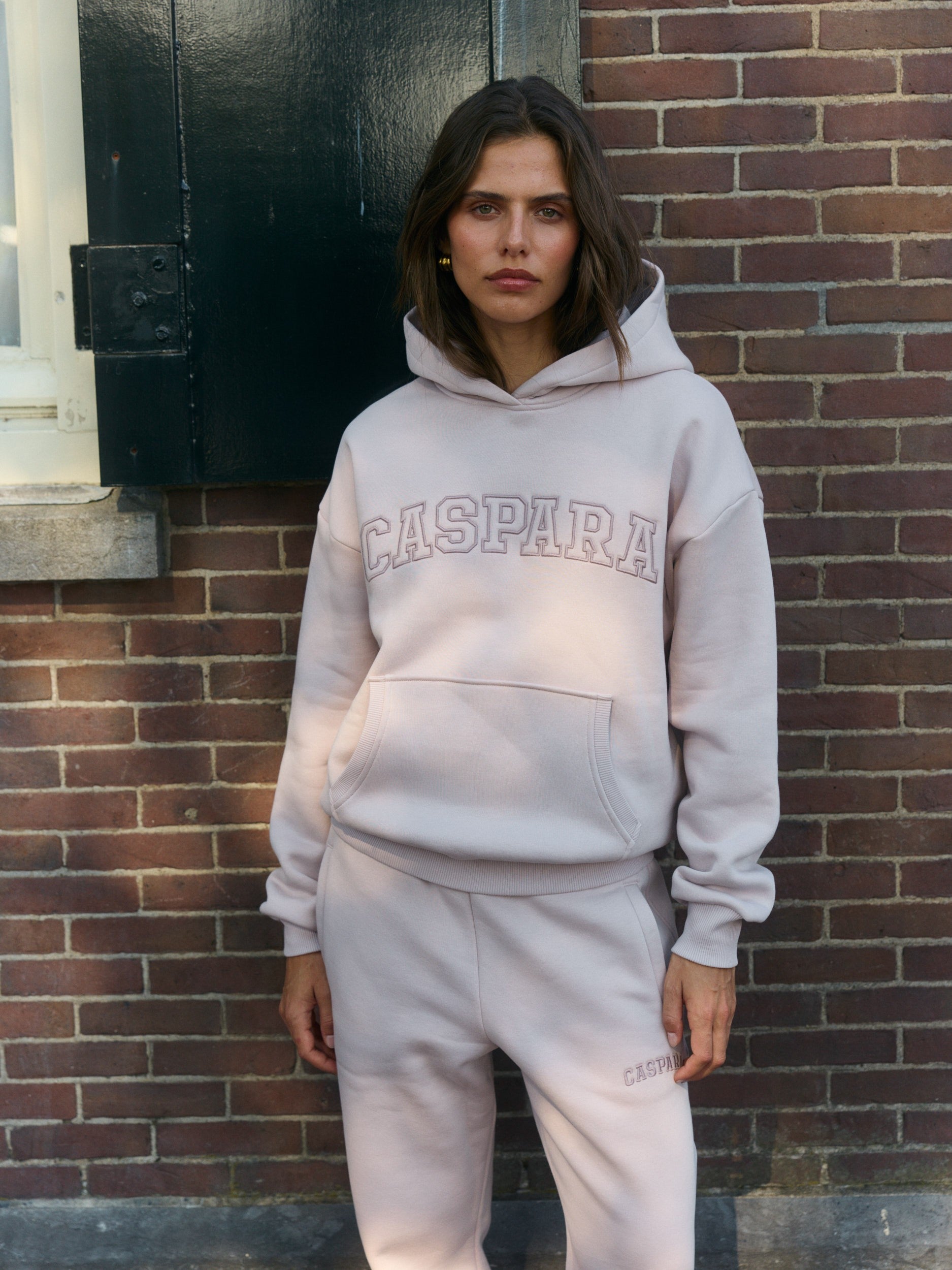 Woman wearing a dusty pink CASPARA hoodie and matching sweatpants against a brick wall.