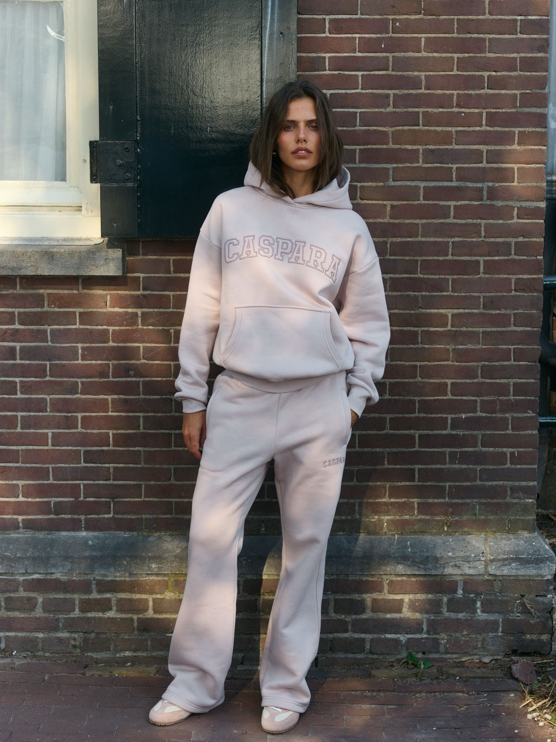 Woman wearing a dusty pink hoodie and matching straight-leg sweatpants against a brick wall.