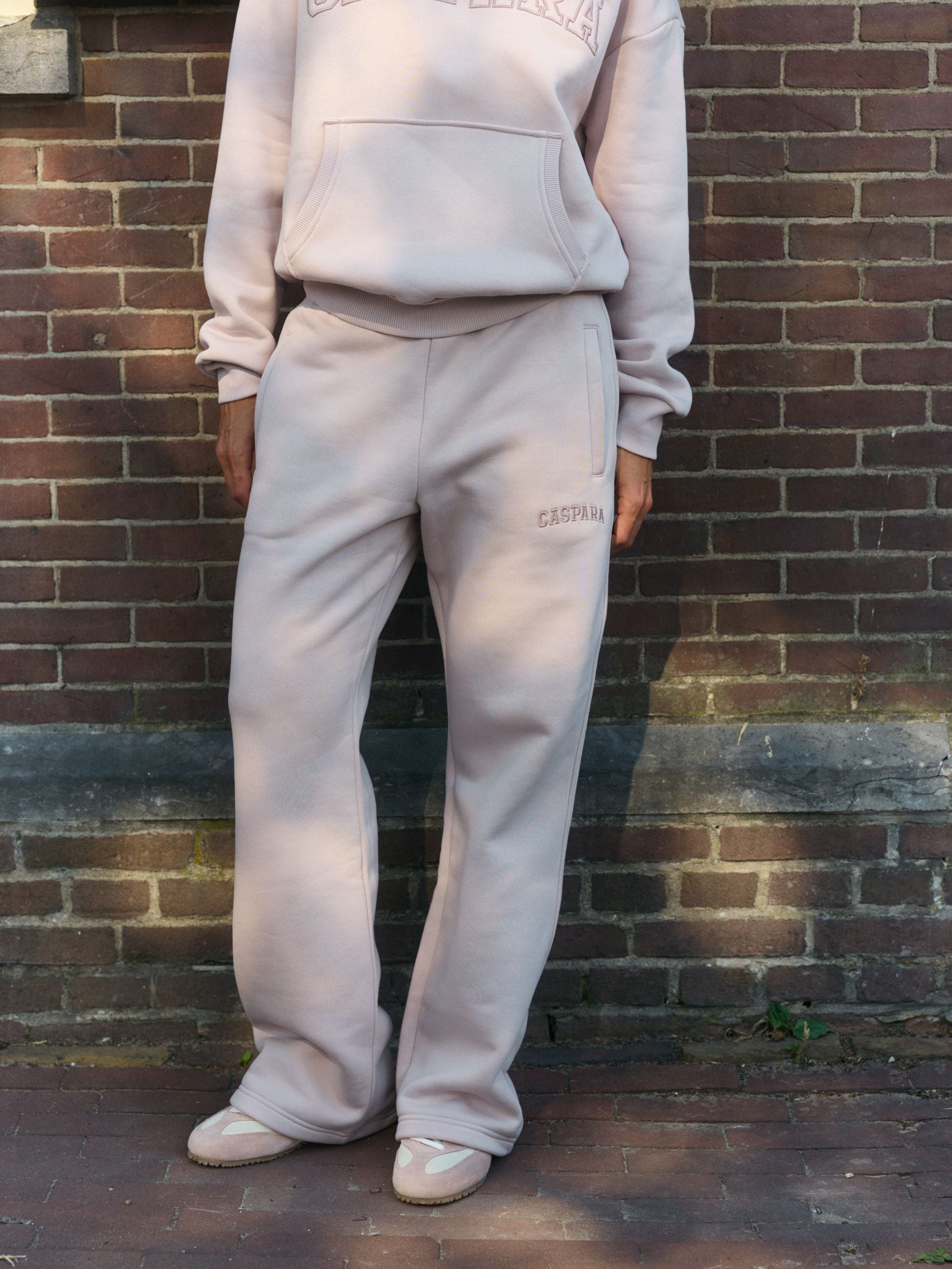 Woman wearing dusty pink straight-leg sweatpants and matching hoodie standing against a brick wall.