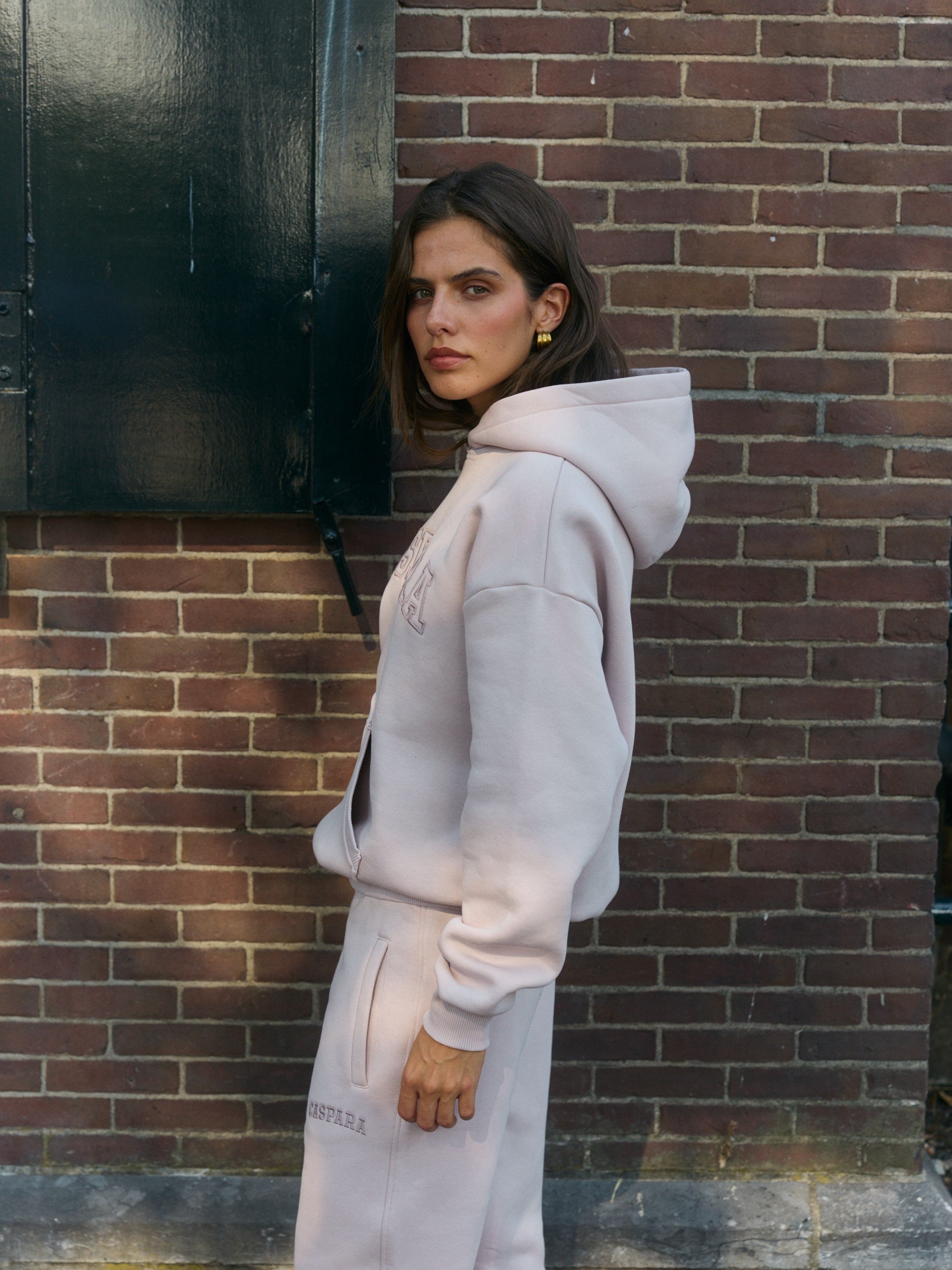 Woman wearing a dusty pink hoodie and matching sweatpants against a brick wall.