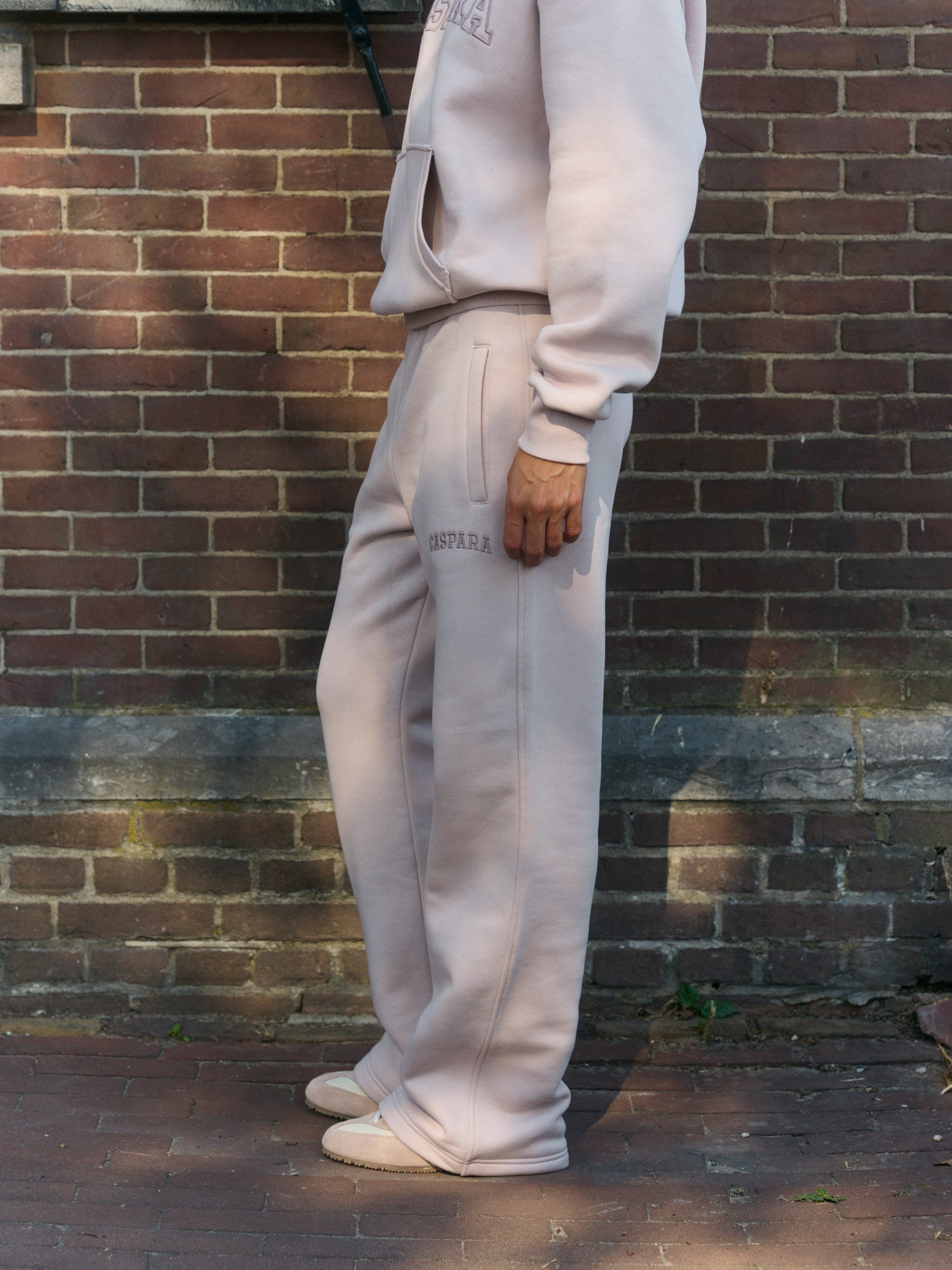 Woman wearing dusty pink straight-leg sweatpants and matching hoodie standing against a brick wall.