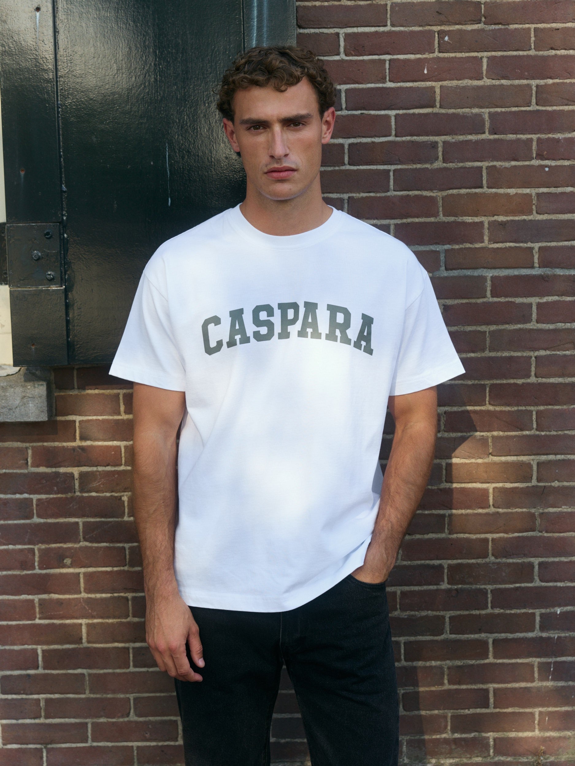 Man wearing a white CASPARA graphic t-shirt standing against a brick wall.