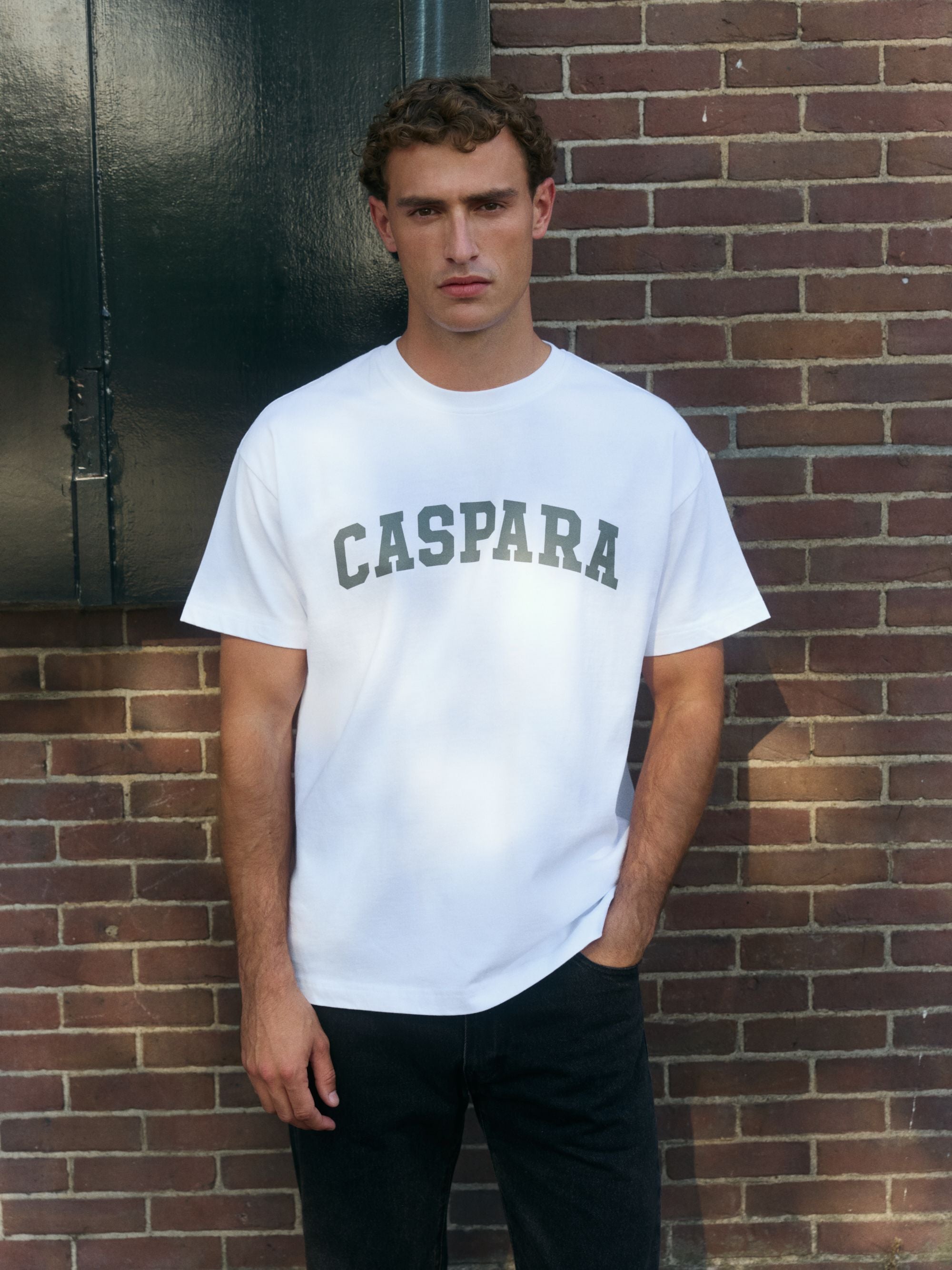 Man wearing a white CASPARA logo t-shirt standing in front of a brick wall.