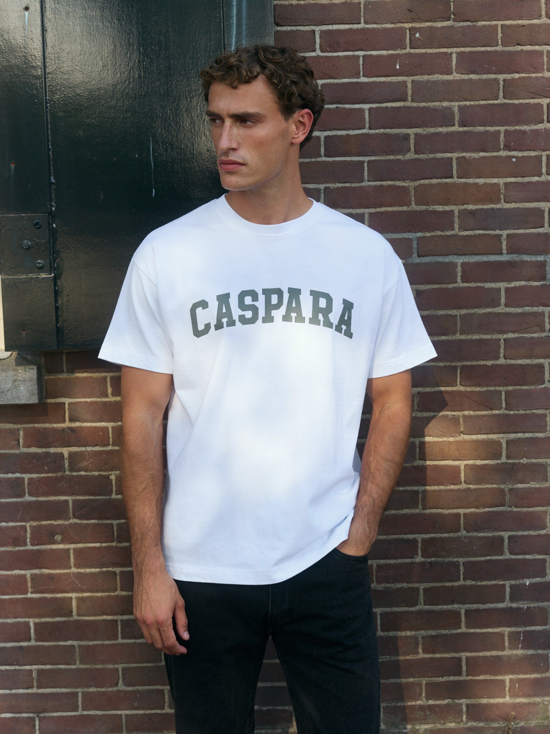 Man wearing a white oversized t-shirt with khaki logo print against a brick wall.