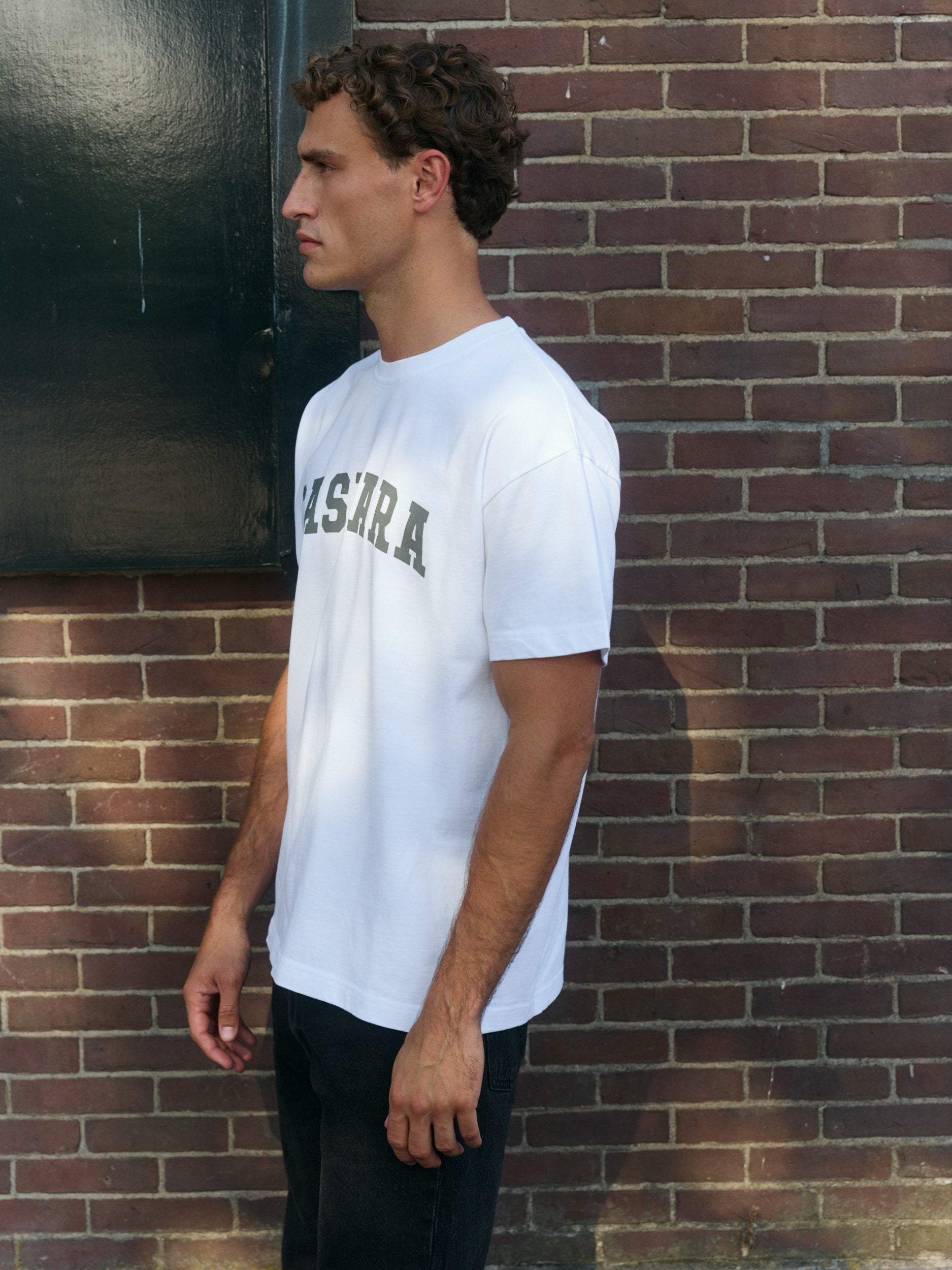 Man wearing a white oversized logo t-shirt standing against a brick wall.
