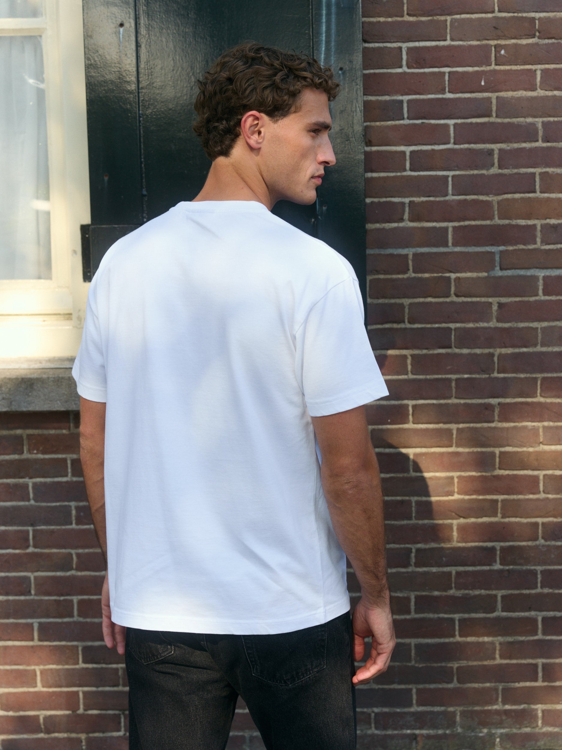 Back view of a man wearing a white oversized cotton t-shirt against a brick wall.