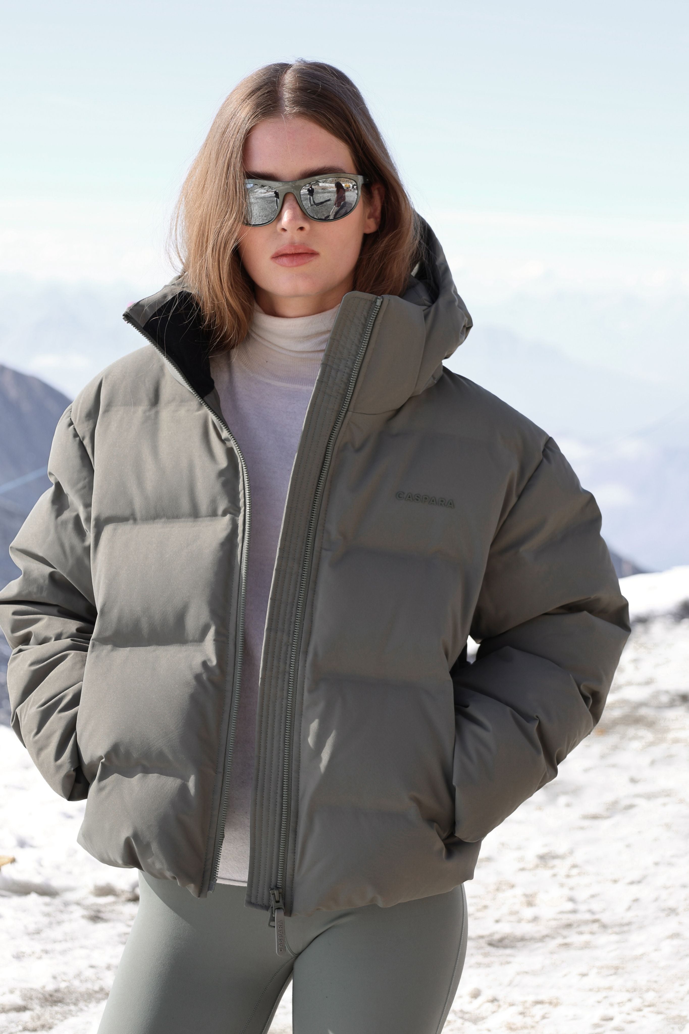 Woman wearing a khaki CASPARA Aurora puffer jacket in snowy mountains.
