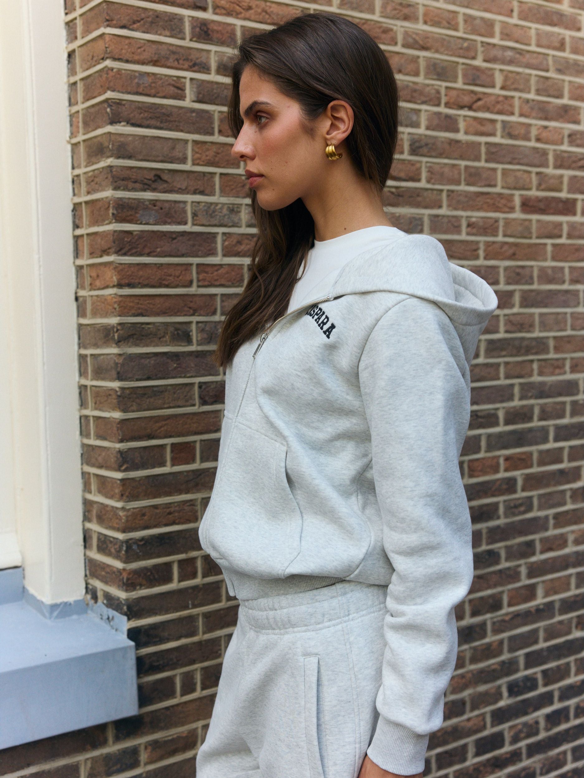 Woman wearing a grey melange zip hoodie and matching sweatpants against a brick wall.