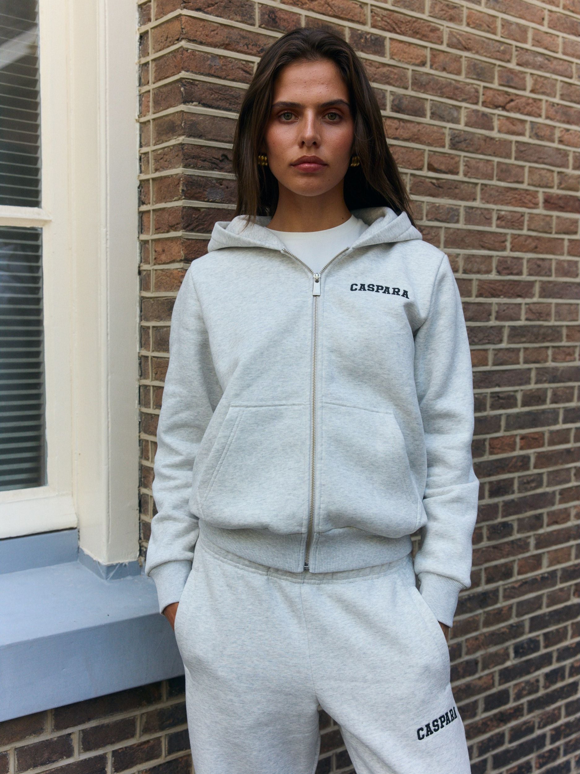 Woman wearing a grey melange zip hoodie and matching sweatpants standing by a brick wall.