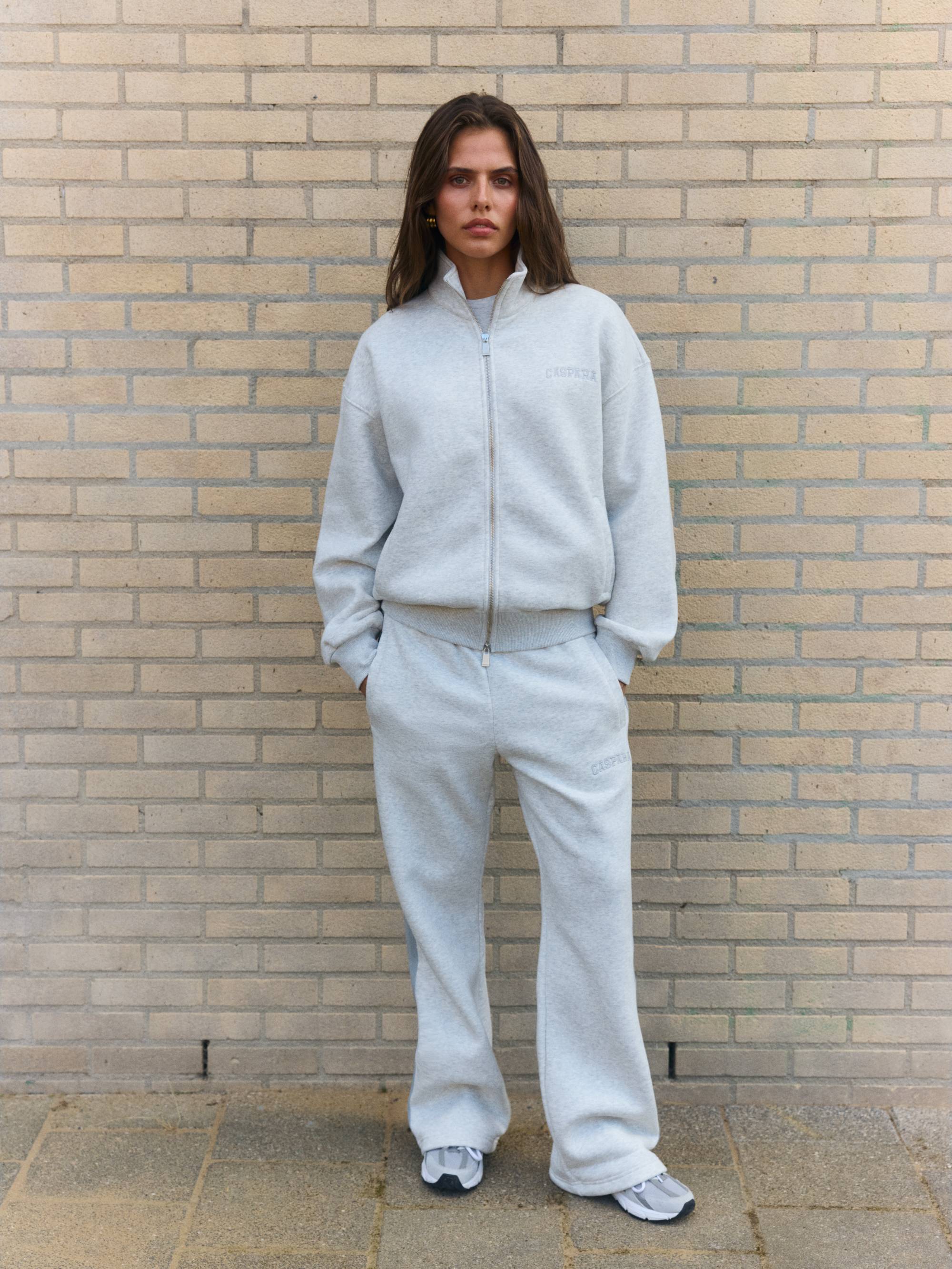 Woman wearing a grey melange CASPARA zip jacket and sweatpants against a brick wall.