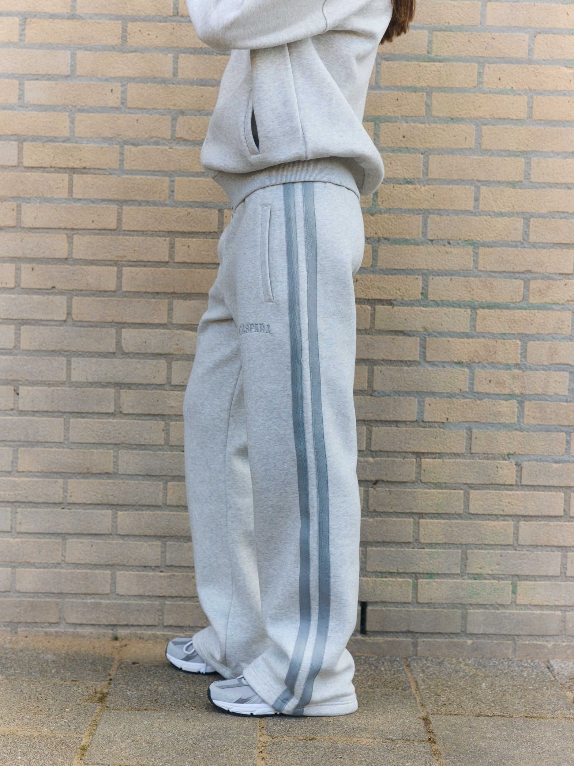 Woman wearing grey melange straight-leg sweatpants with side stripes against a brick wall.