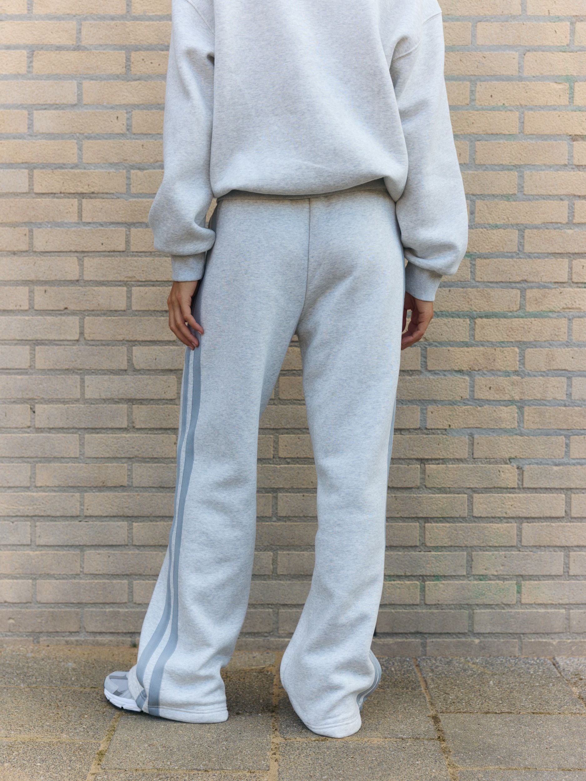 Woman wearing grey melange straight-leg sweatpants with side stripes against a brick wall.