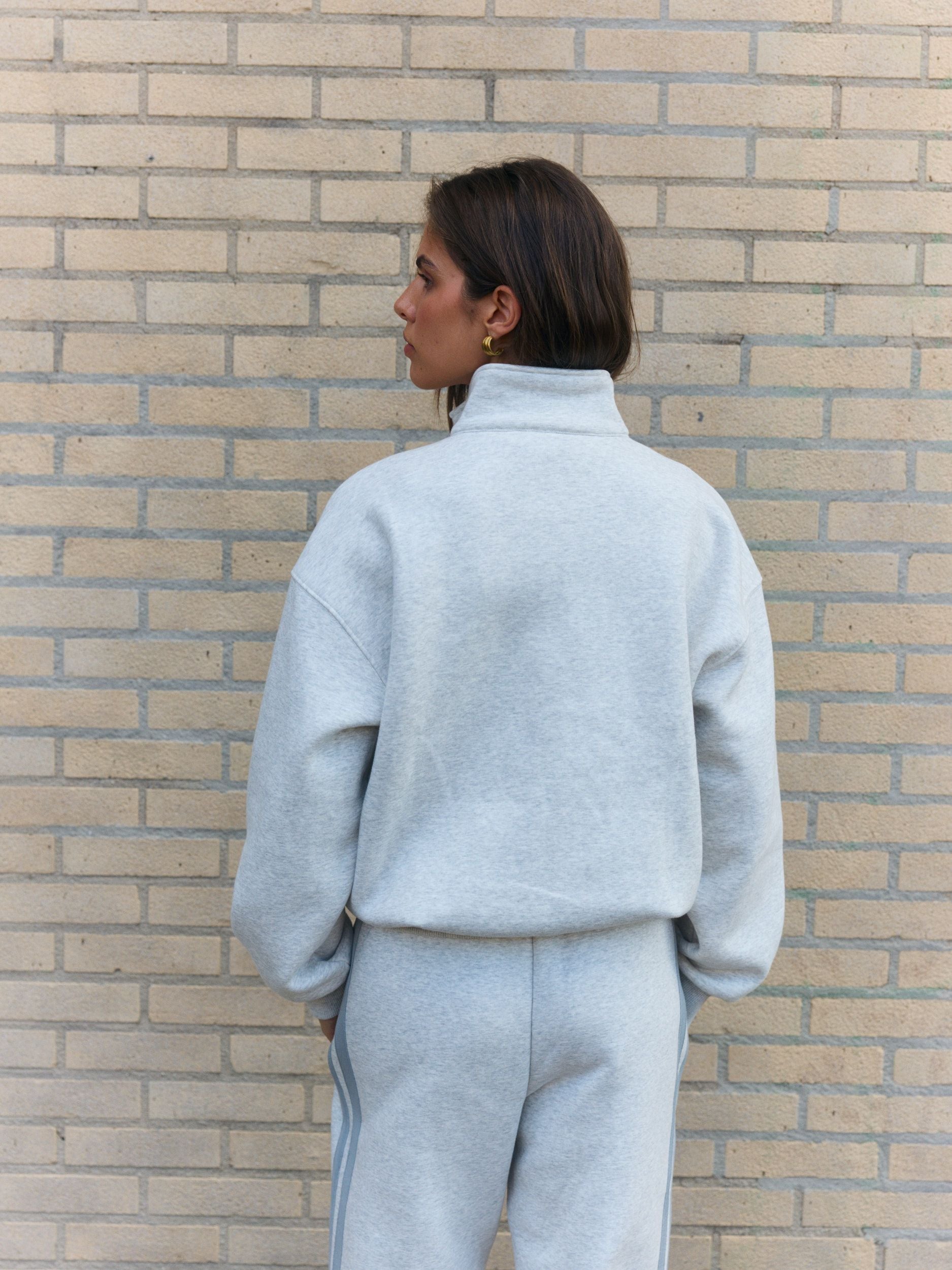 Woman wearing a grey melange funnel neck jacket and sweatpants against a brick wall.