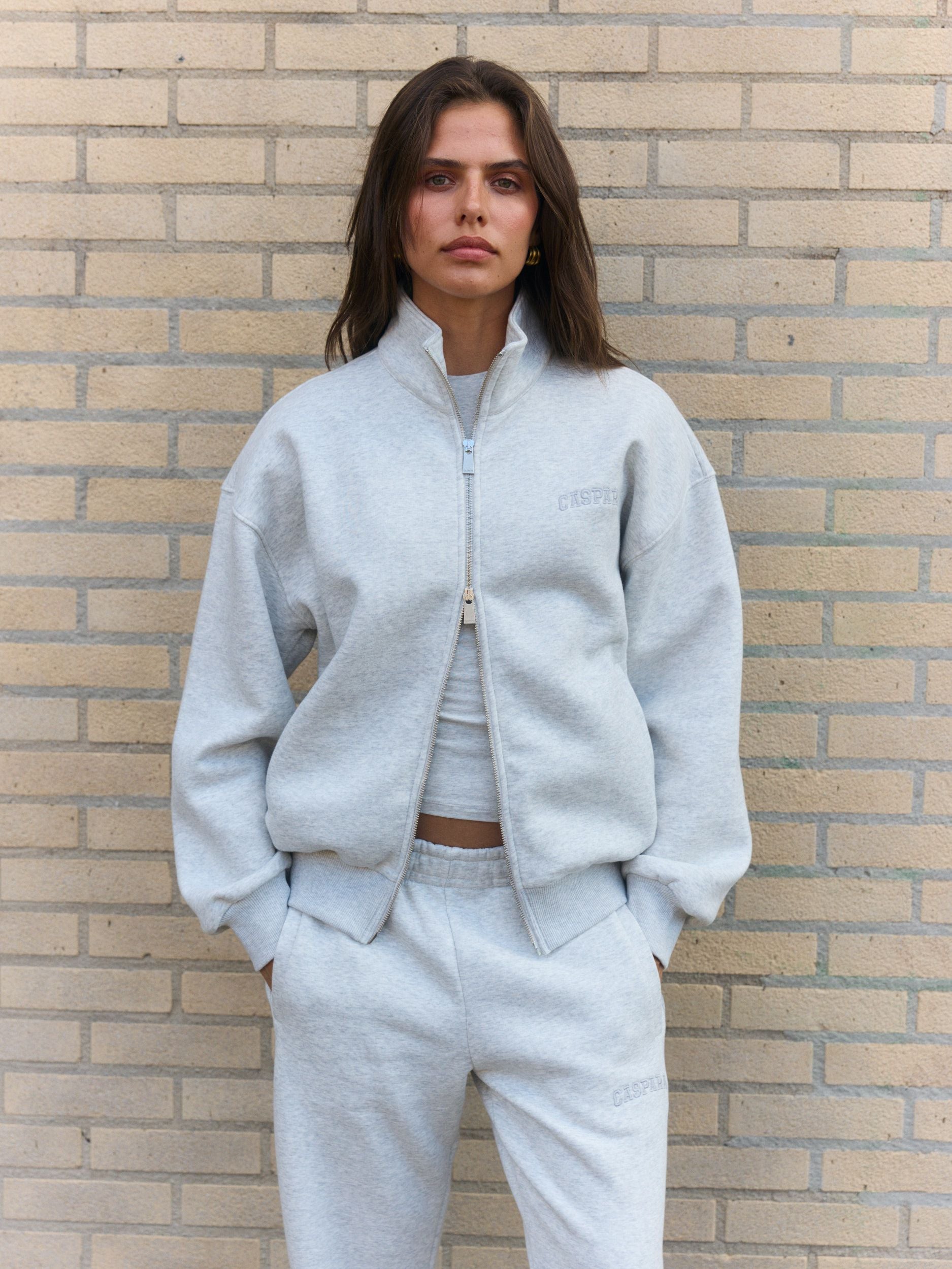 Woman wearing a grey melange funnel neck zip jacket against a brick wall.
