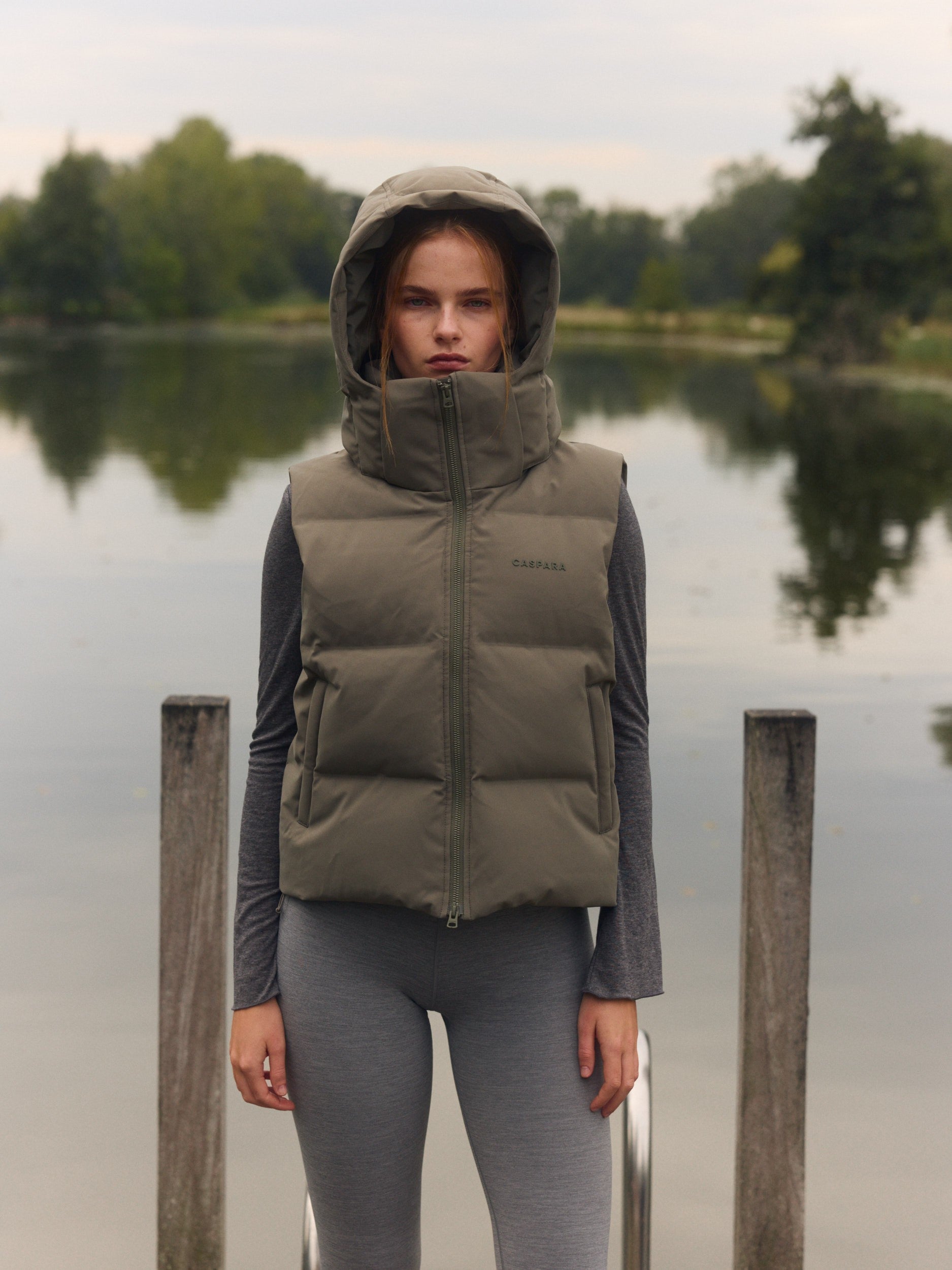 Woman wearing a khaki hooded puffer vest and grey leggings standing outdoors by a lake.