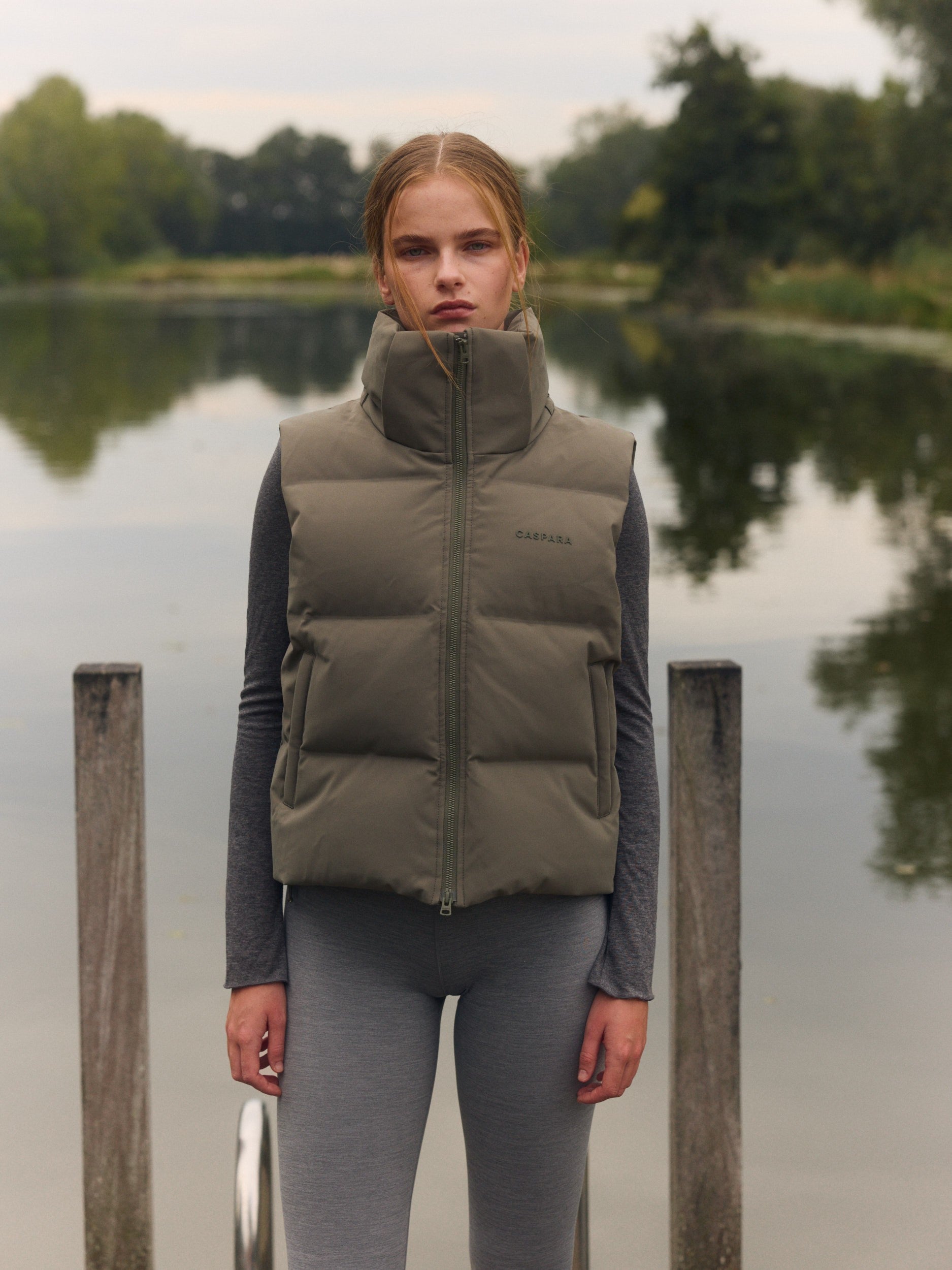 Woman wearing a khaki cropped puffer vest with a high neck standing on a dock by a lake.