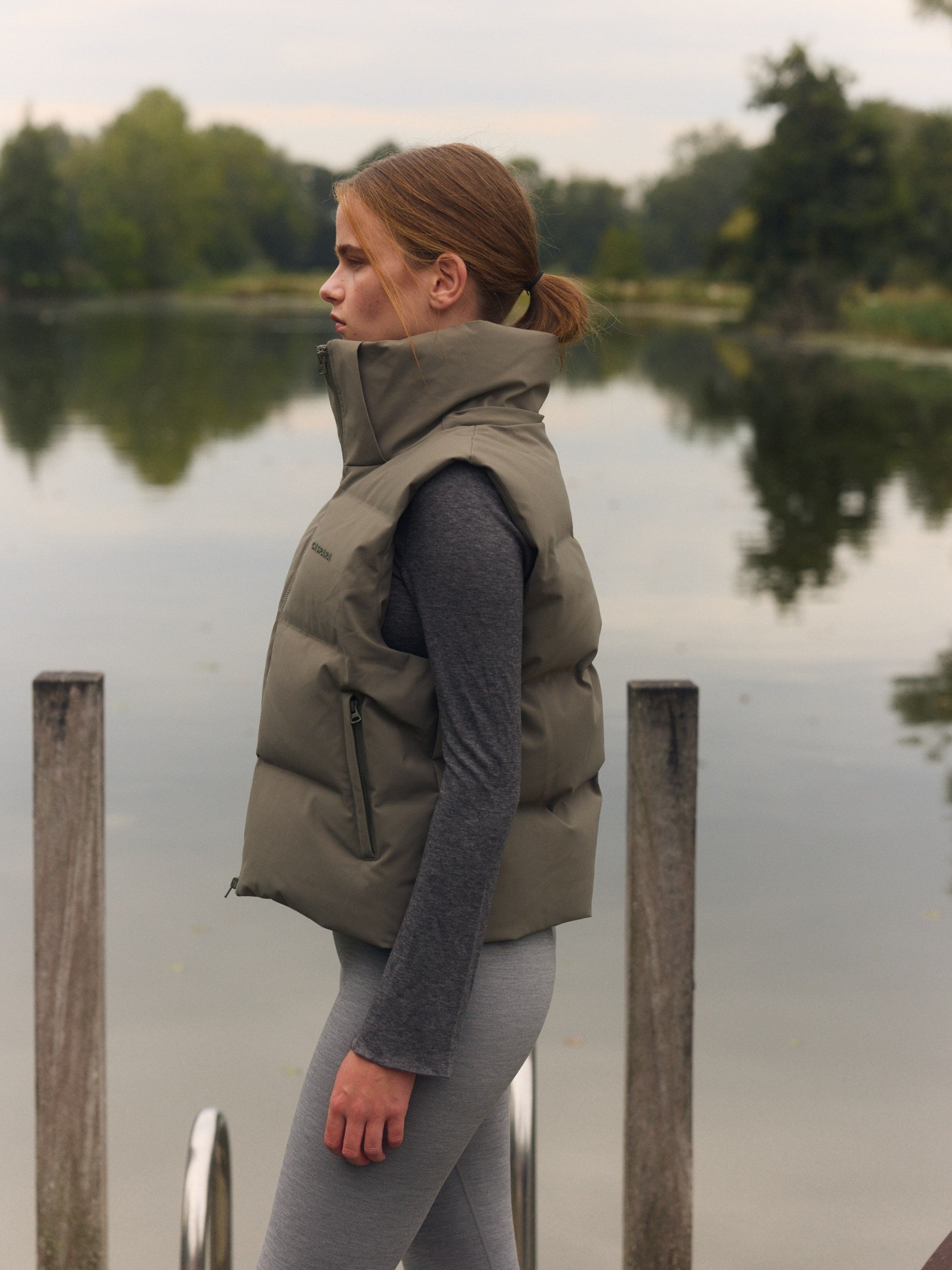 Woman wearing a khaki puffer vest with a high collar standing outdoors by a lake.