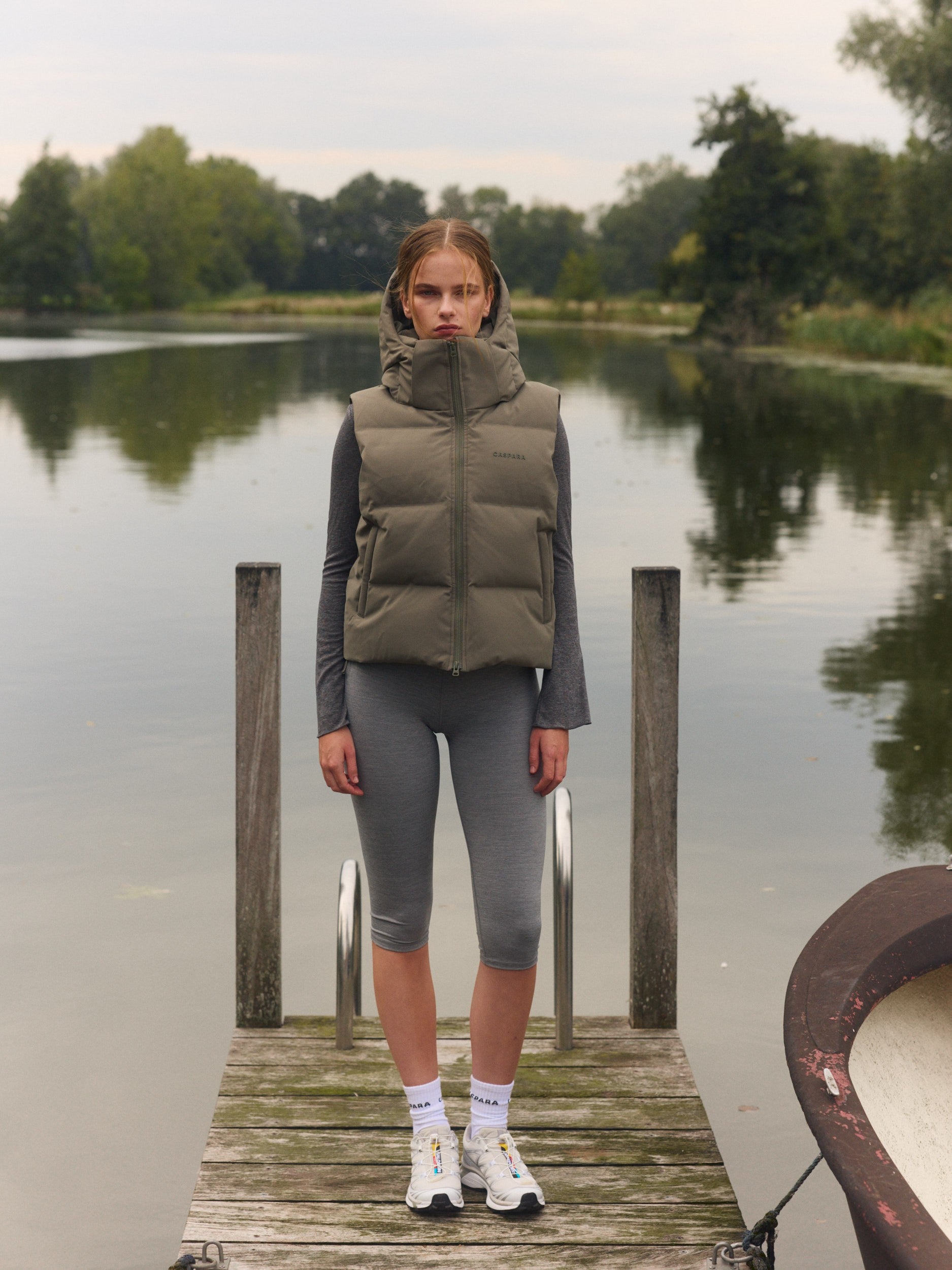 Woman wearing a khaki hooded puffer vest and grey tights standing on a wooden dock by a lake.