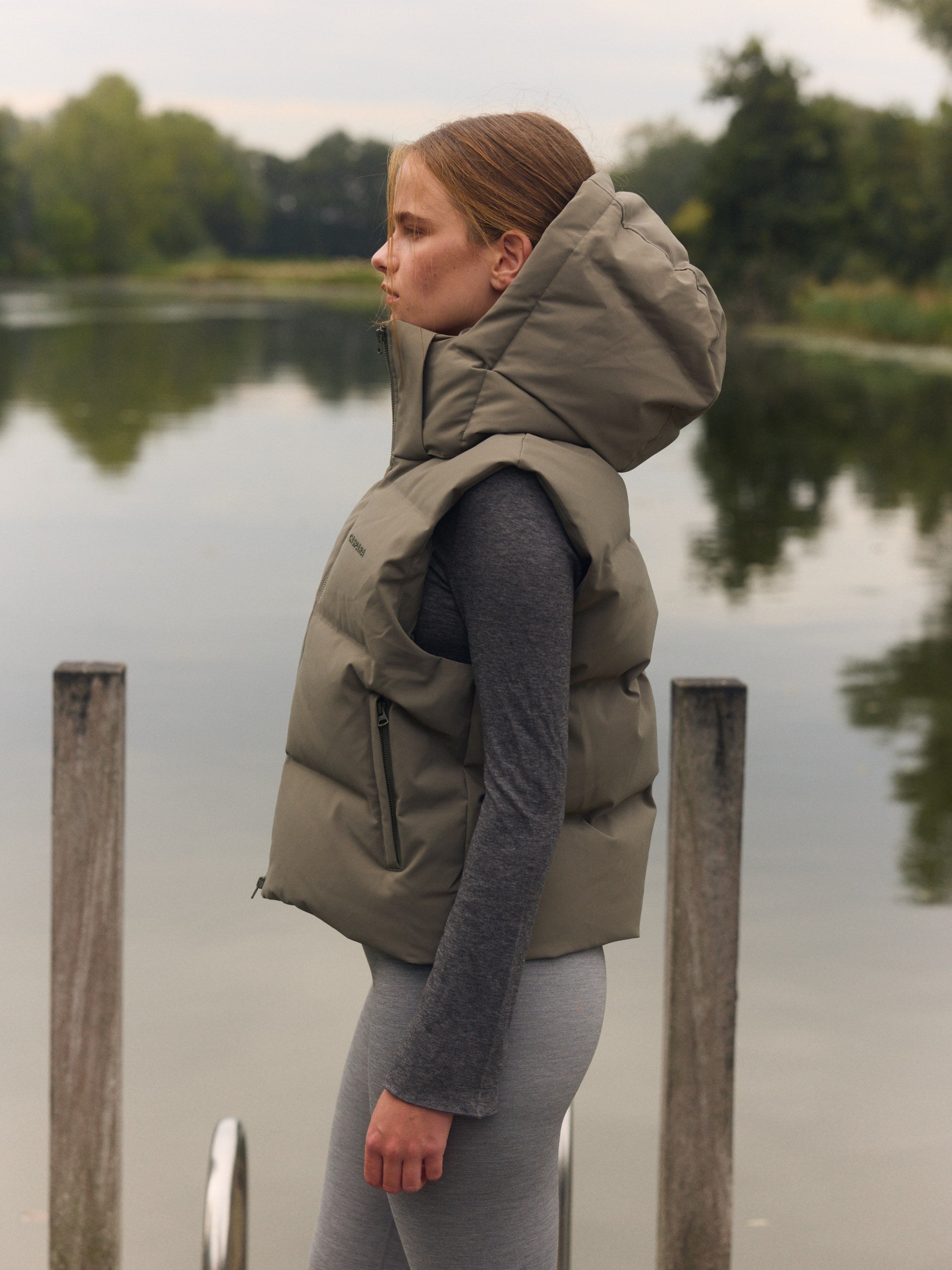 Woman wearing a khaki hooded puffer vest standing outdoors beside a lake.